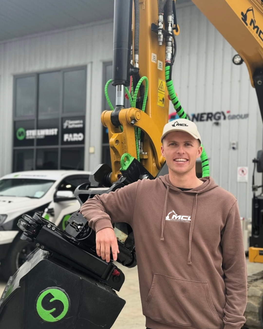A young man standing outdoors next to construction equipment and a white car, wearing a beige cap and a brown hoodie with a logo, smiling at the camera.