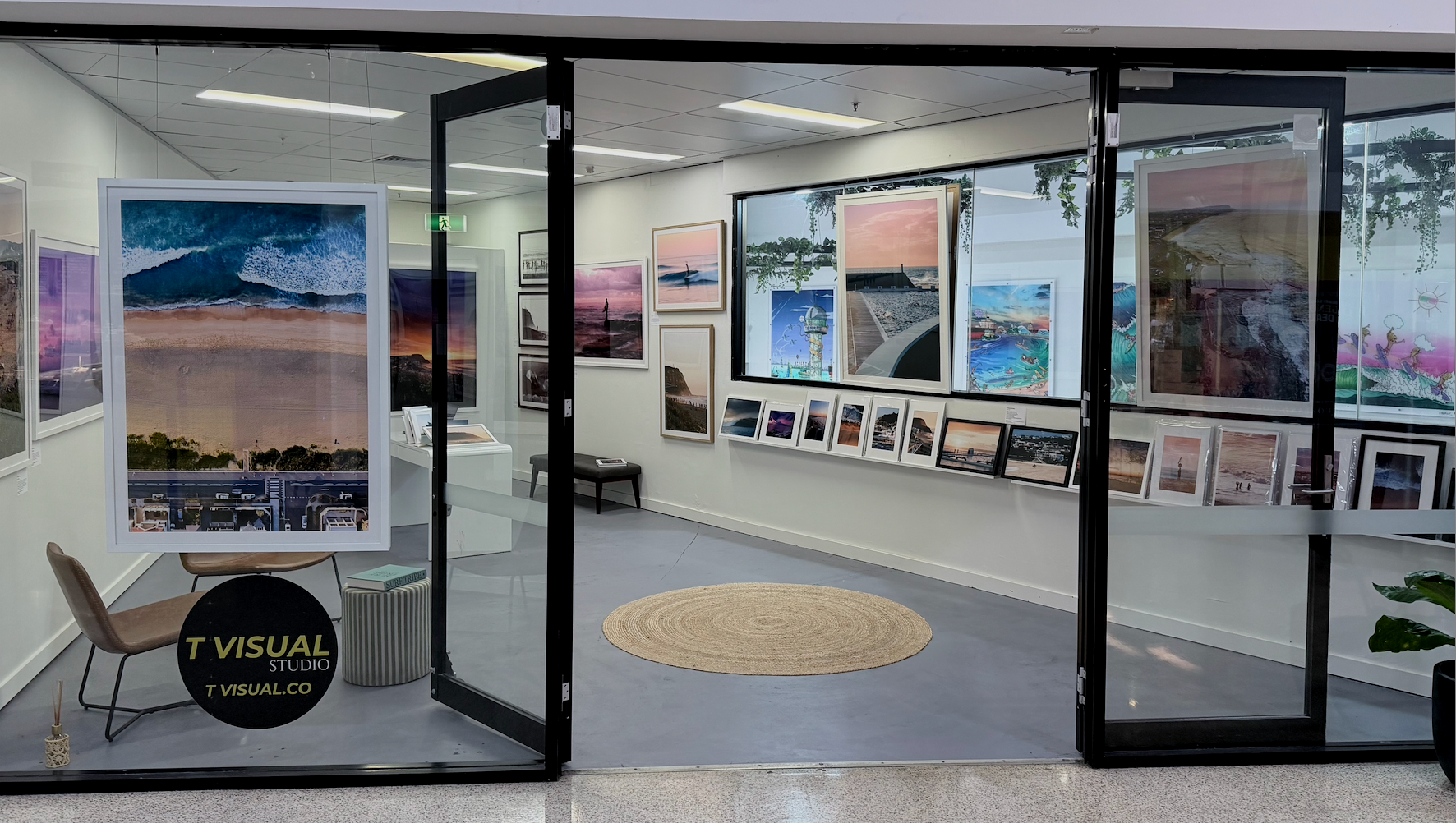 Interior of an art gallery with framed photographs of landscapes on white walls, a central display table with smaller framed images, and seating. The gallery has a light wood floor and ceiling lights.