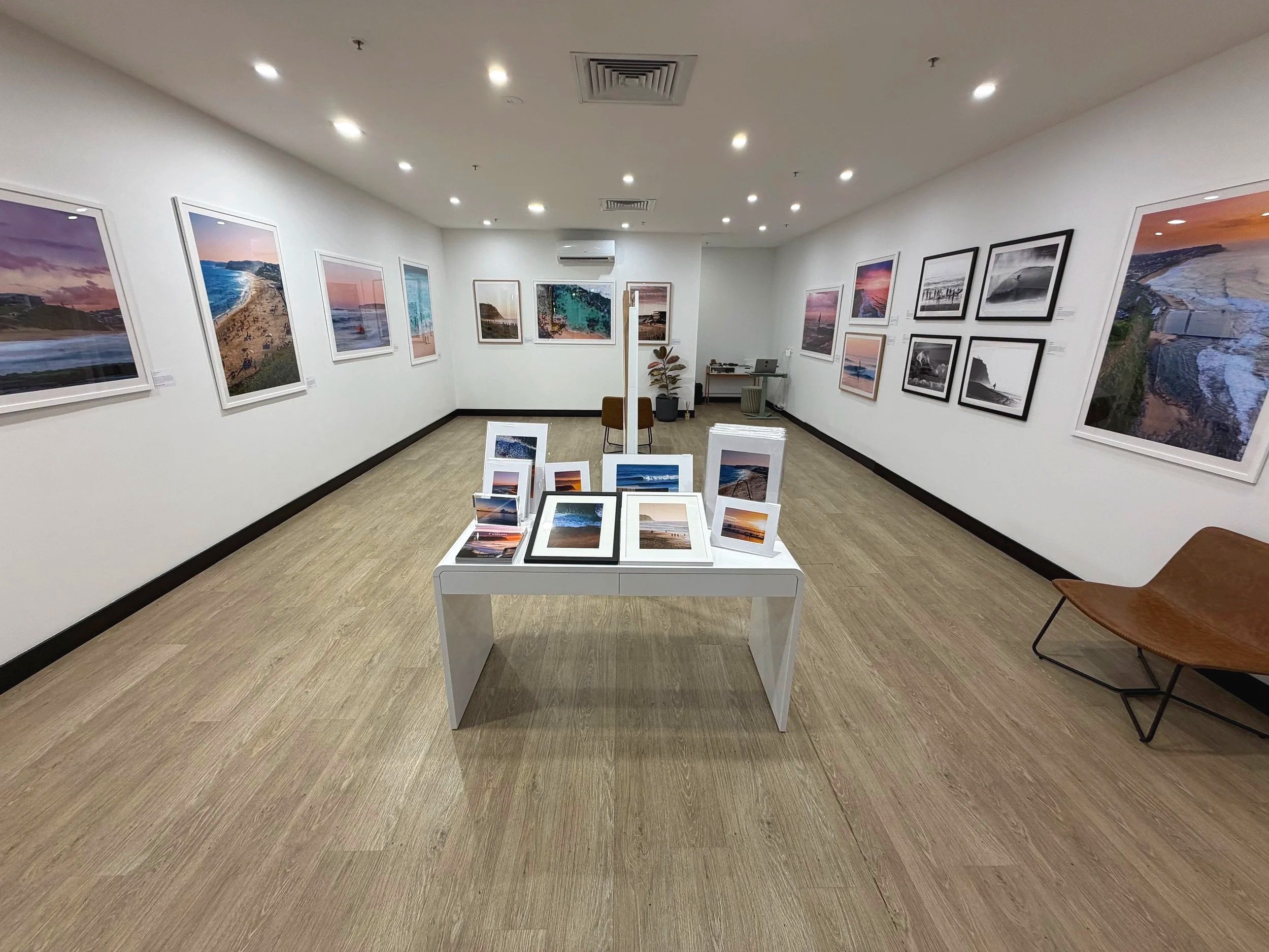Interior of an art gallery with framed photographs of landscapes on white walls, a central display table with smaller framed images, and seating. The gallery has a light wood floor and ceiling lights.