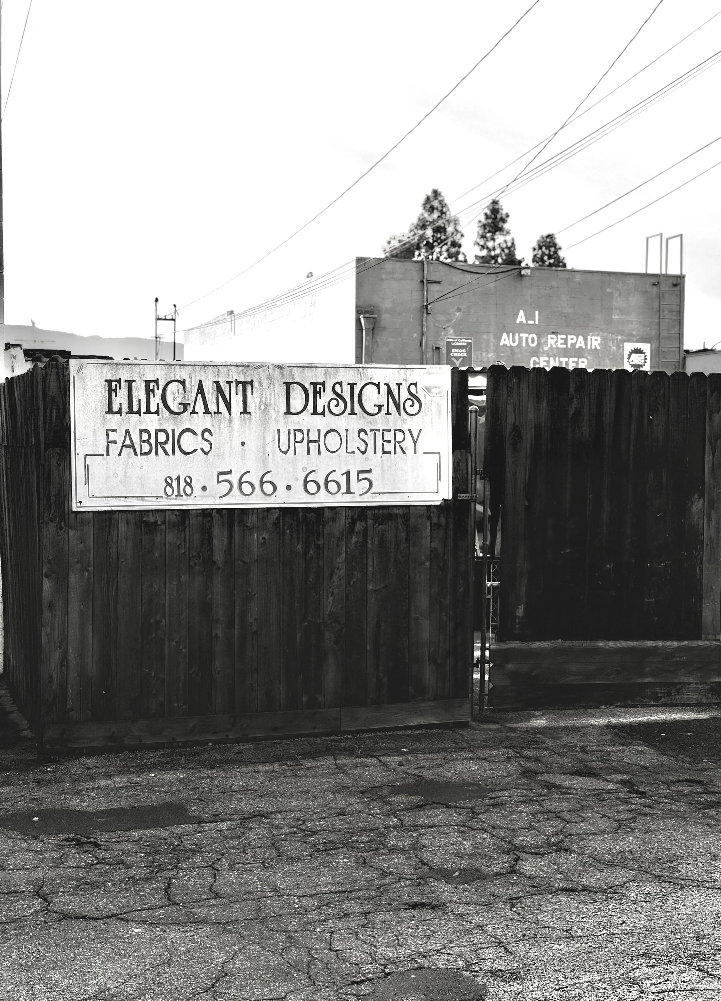 Black and white photo of a wooden fence with a sign for Elegant Designs Fabrics and Upholstery, along with a phone number. Behind the fence, a building with the sign A.I Auto Repair Center is visible in the background.