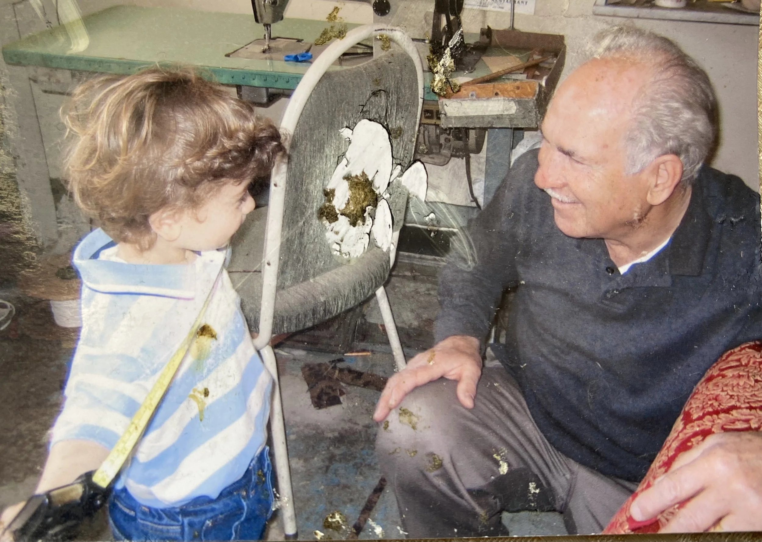 A young boy and an older man sharing a joyful moment in a workshop, with the boy holding a measuring tape and the man smiling at him. The background features a workbench with tools and safety gloves.