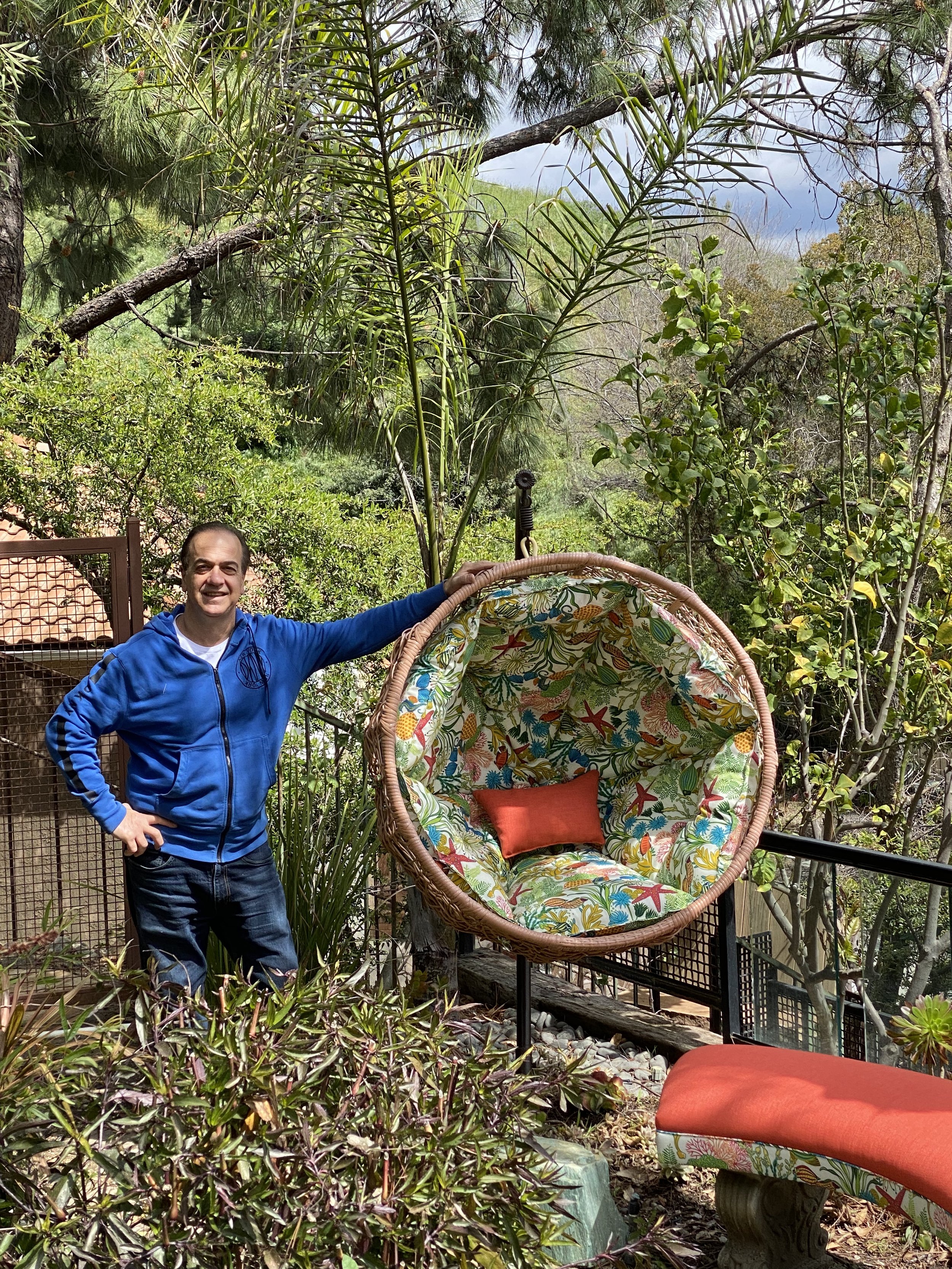 A man in a blue jacket with a big smile standing next to a hanging papasan chair with colorful floral fabric and a red cushion in a lush garden with trees and bushes.