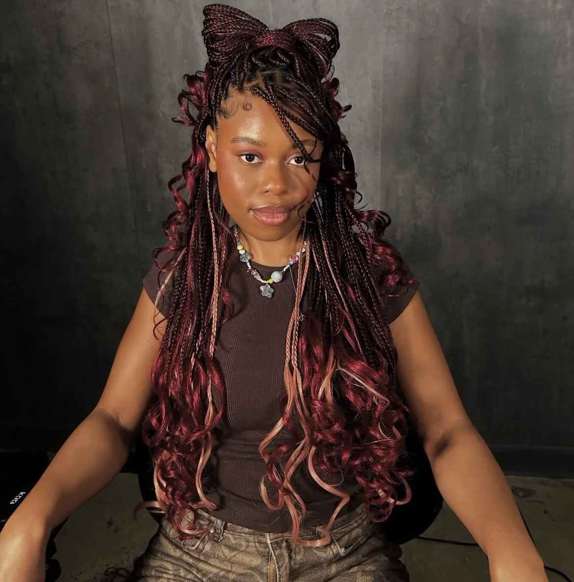 A woman with long, curly, pink and black braids styled with a bow on top, sitting against a dark background, wearing a black top and colorful beaded necklace.