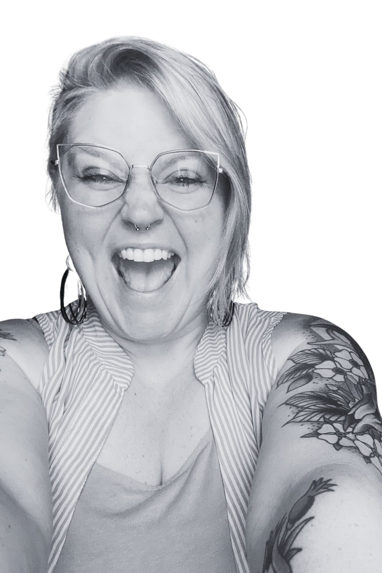 A woman with glasses, tattoos on her arms, and a septum piercing, smiling and sticking out her tongue, taking a selfie with arms extended.