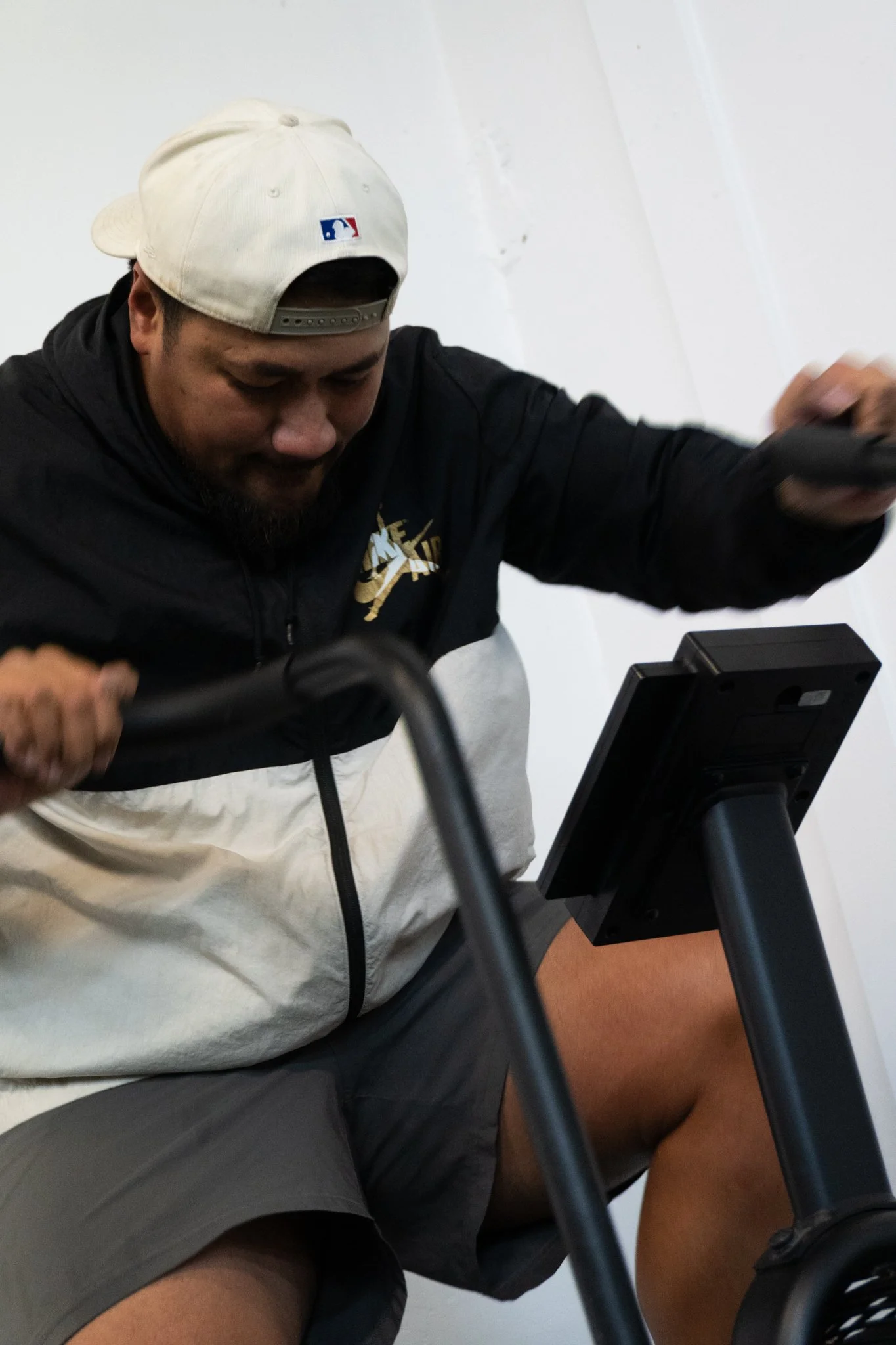 A man wearing a white baseball cap and black Nike jacket is sitting on an exercise bike, looking down and pedaling with effort.