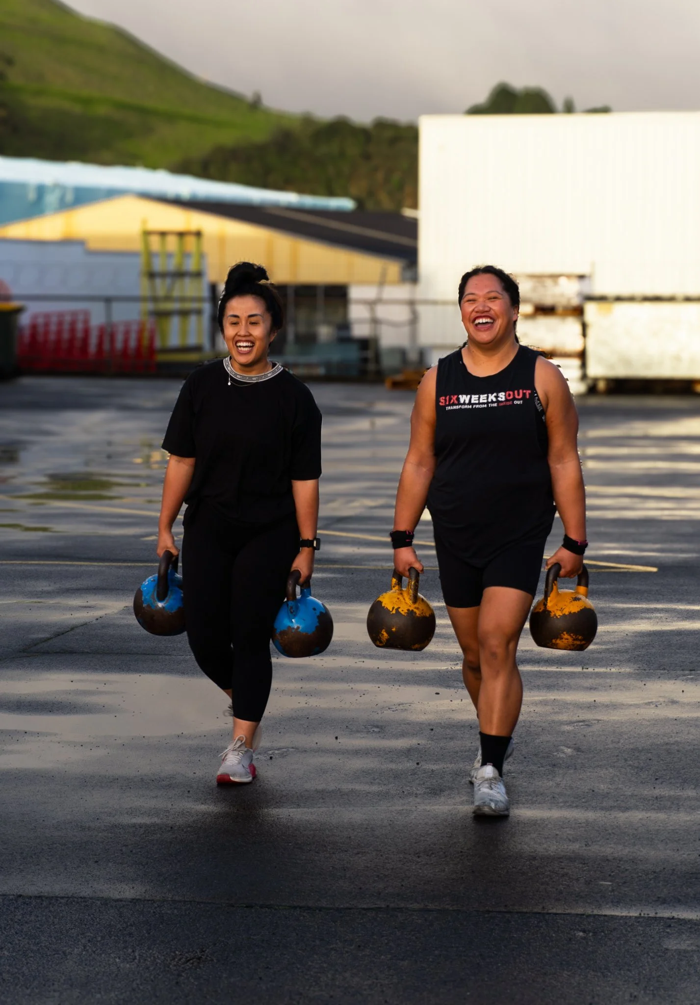 Two women walking on an outdoor surface, carrying kettlebells and smiling, with industrial and mountainous background.