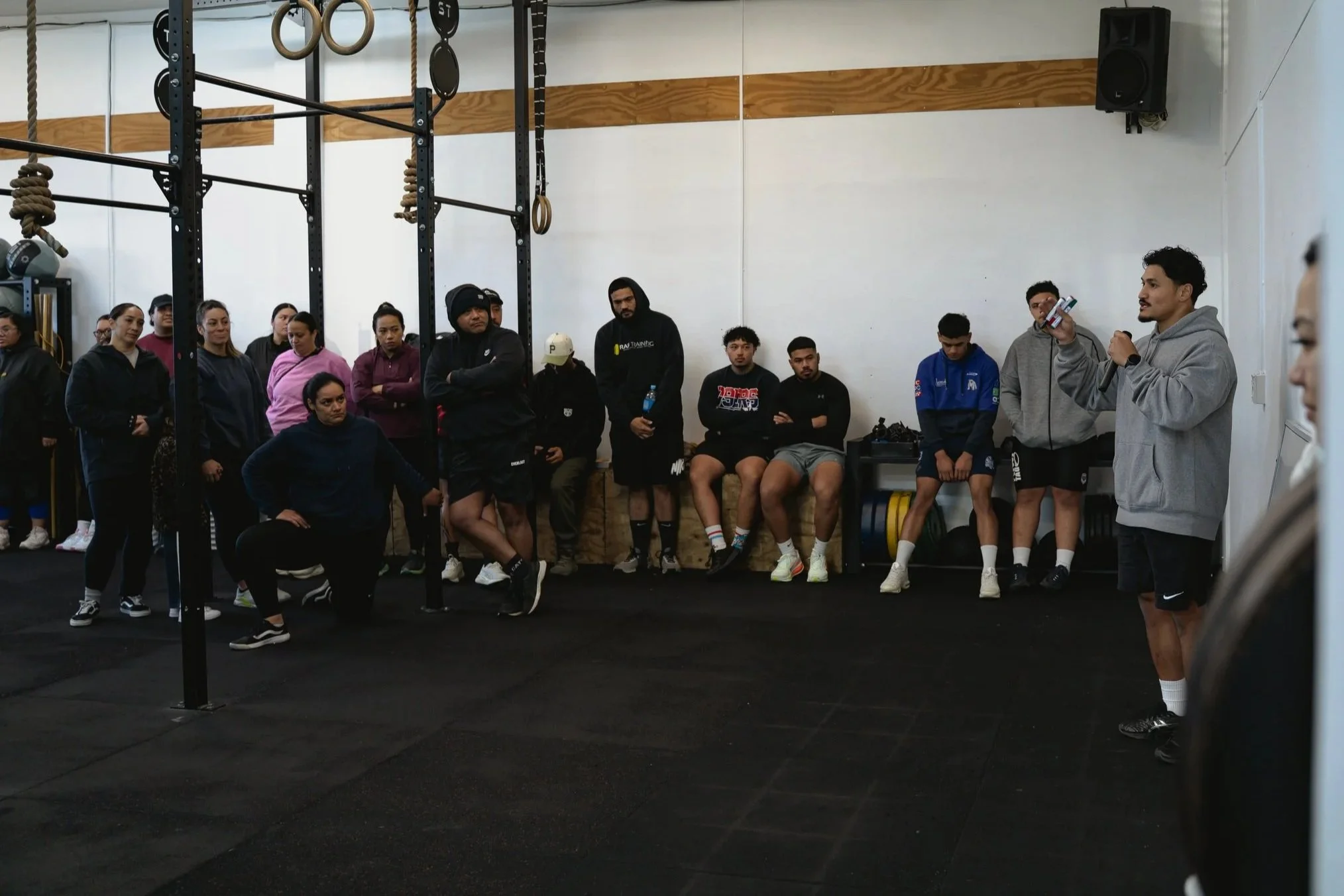 A group of athletes listening to a trainer in a gym. The trainer is speaking into a microphone and holding a small device. The athletes are standing or sitting against the wall, some wearing athletic clothes and sneakers. Gym equipment and climbing r