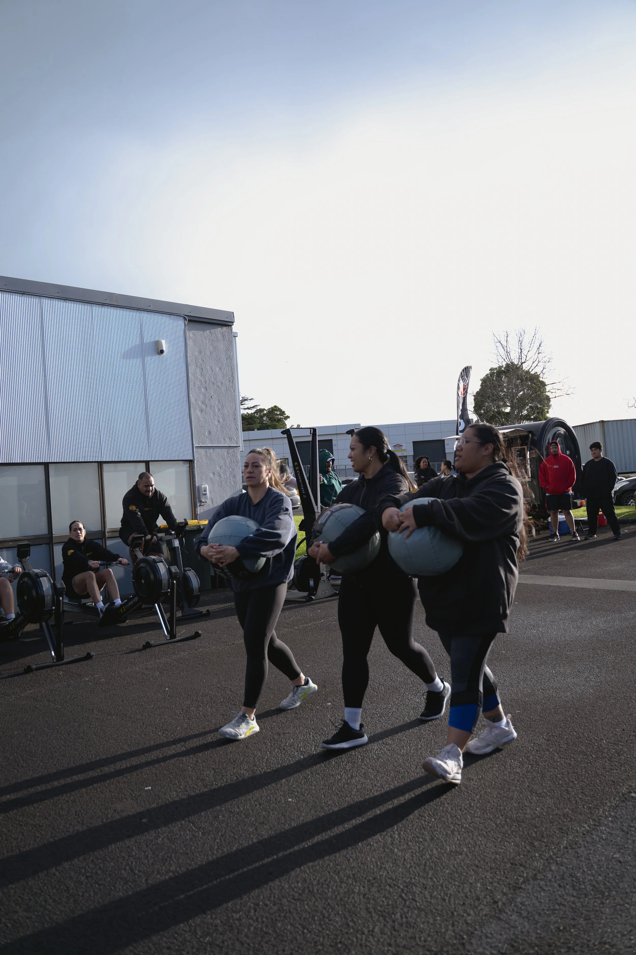 Group of people walking with weight-plate armor outside of a gym, some sitting on rowing machines.