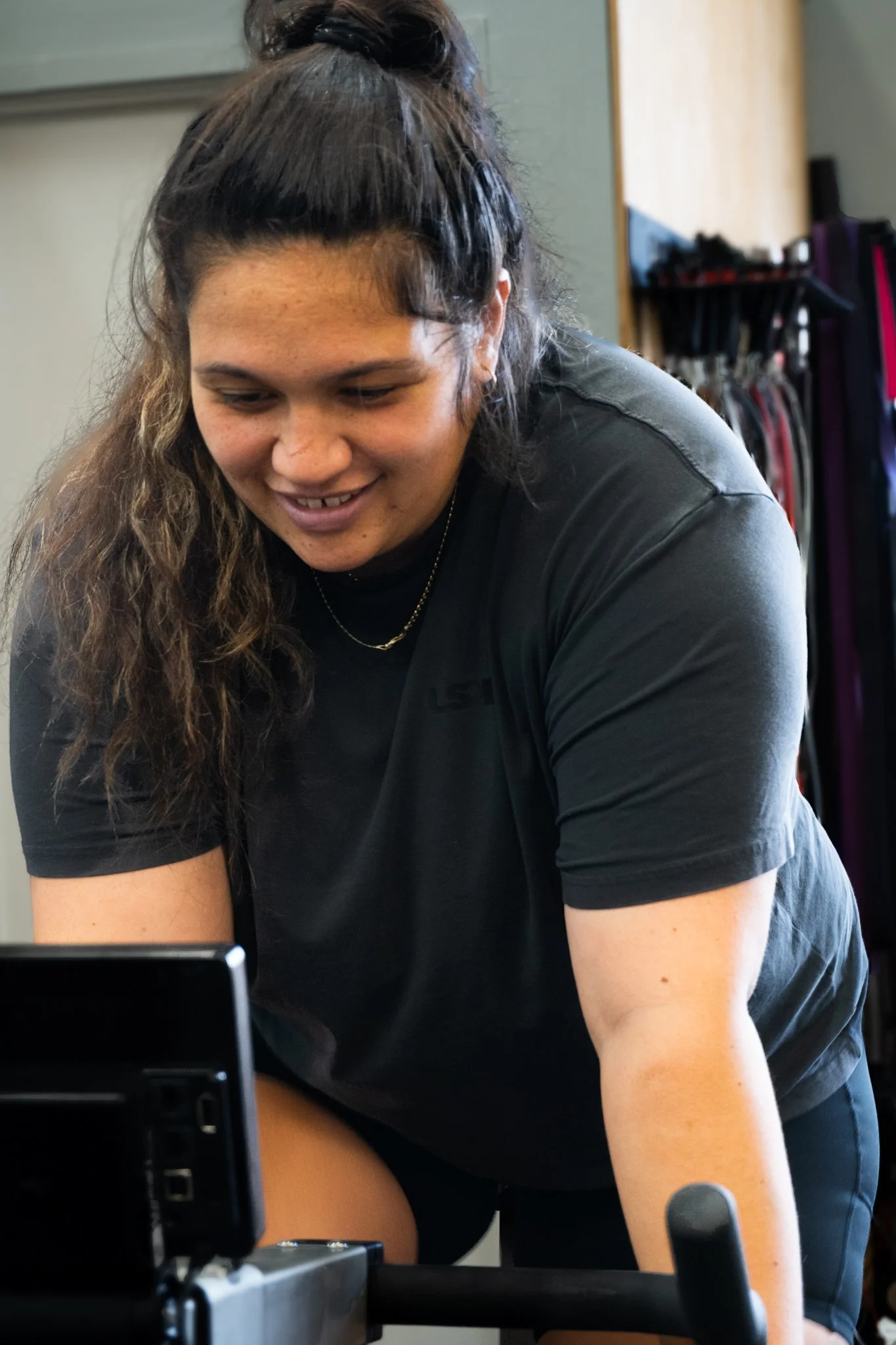 A young woman with curly hair tied in a bun, wearing a black shirt and sitting on a rowing machine, smiling and looking down at a device.