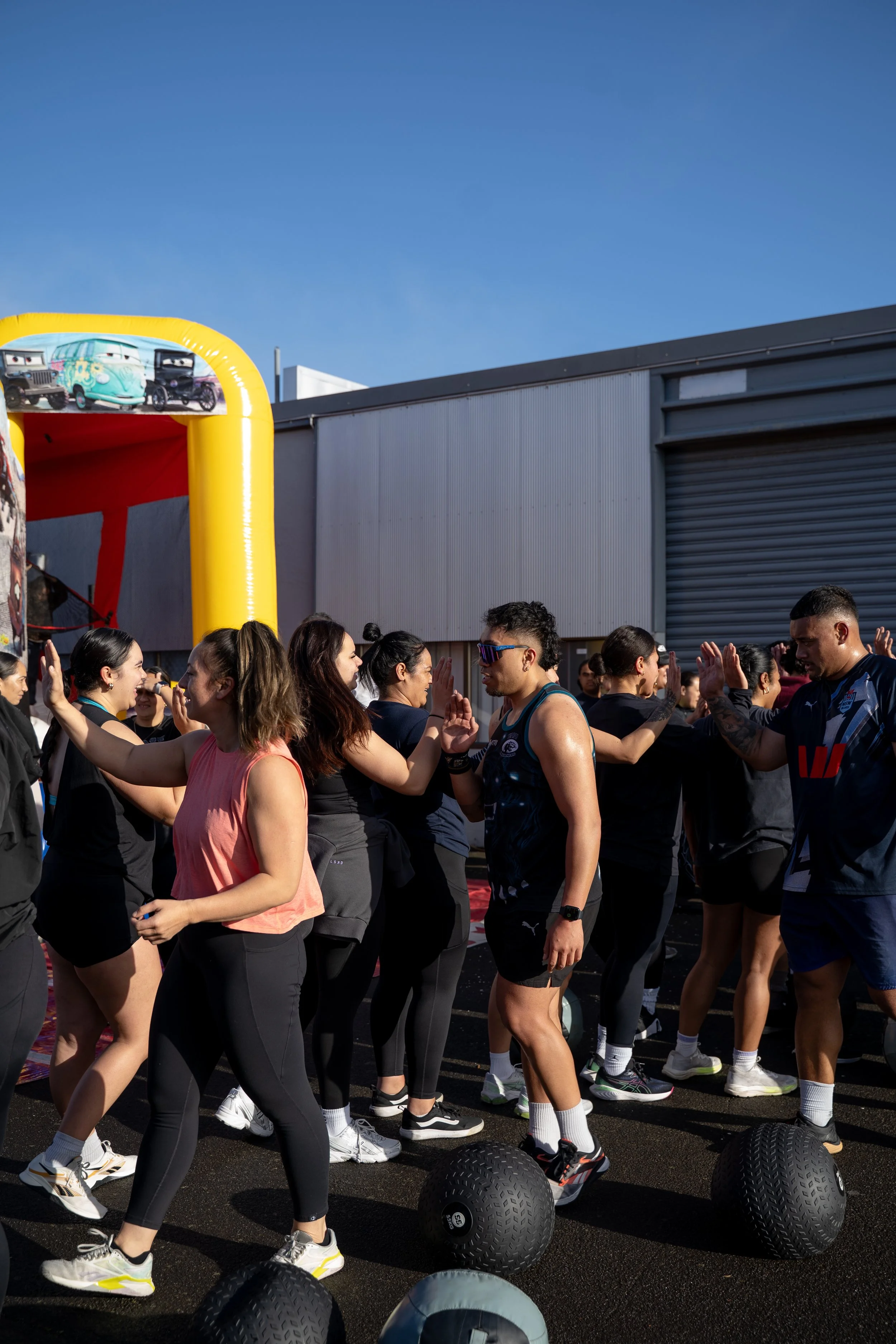 People participating in a group high-five after a workout or exercise session outdoors, with fitness balls on the ground and colorful inflatable arch in the background under a clear blue sky.