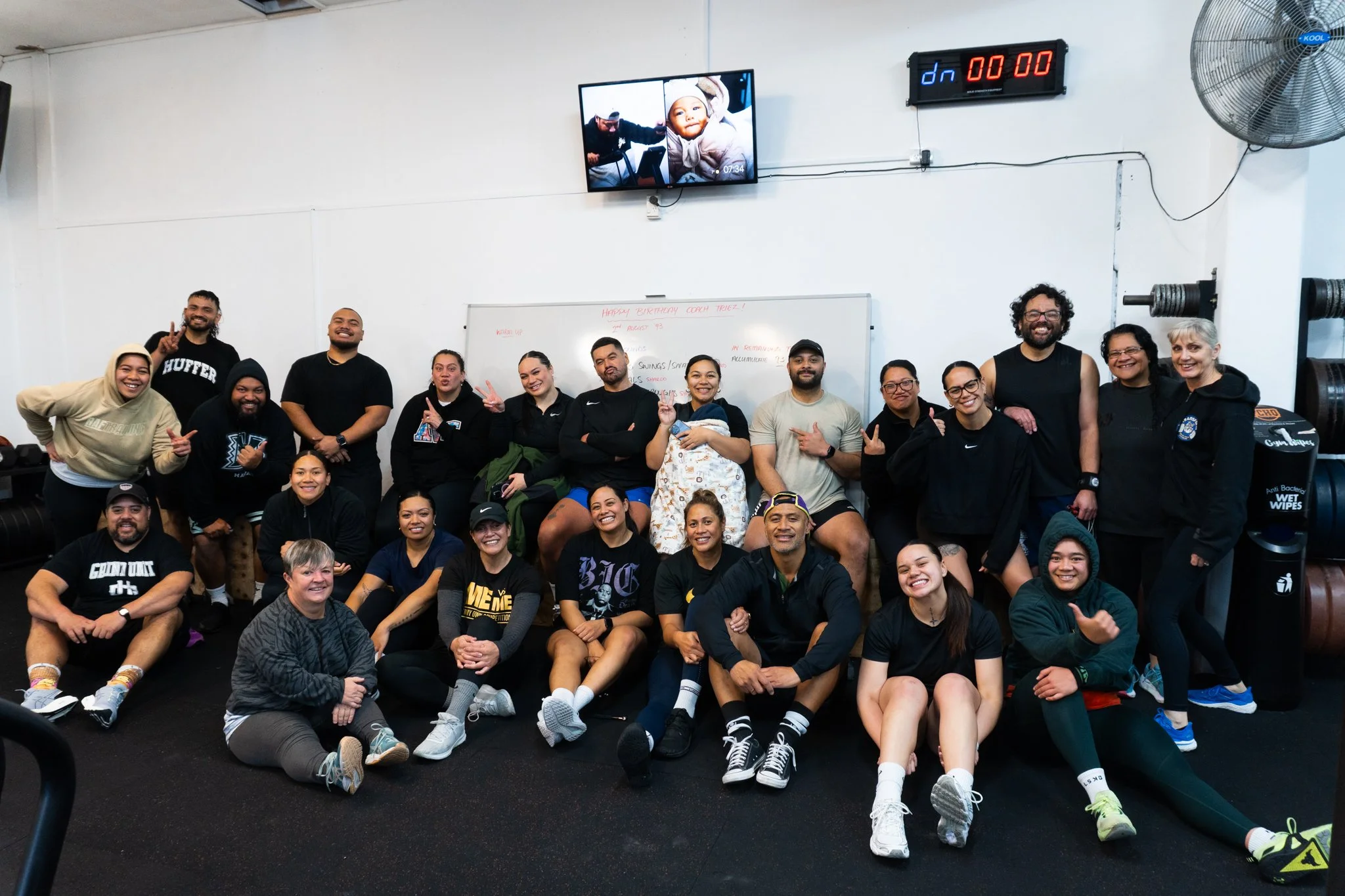 Group of people posing for a photo in a gym, with some sitting on the floor and others standing. The gym has a whiteboard, a clock, and exercise equipment in the background.