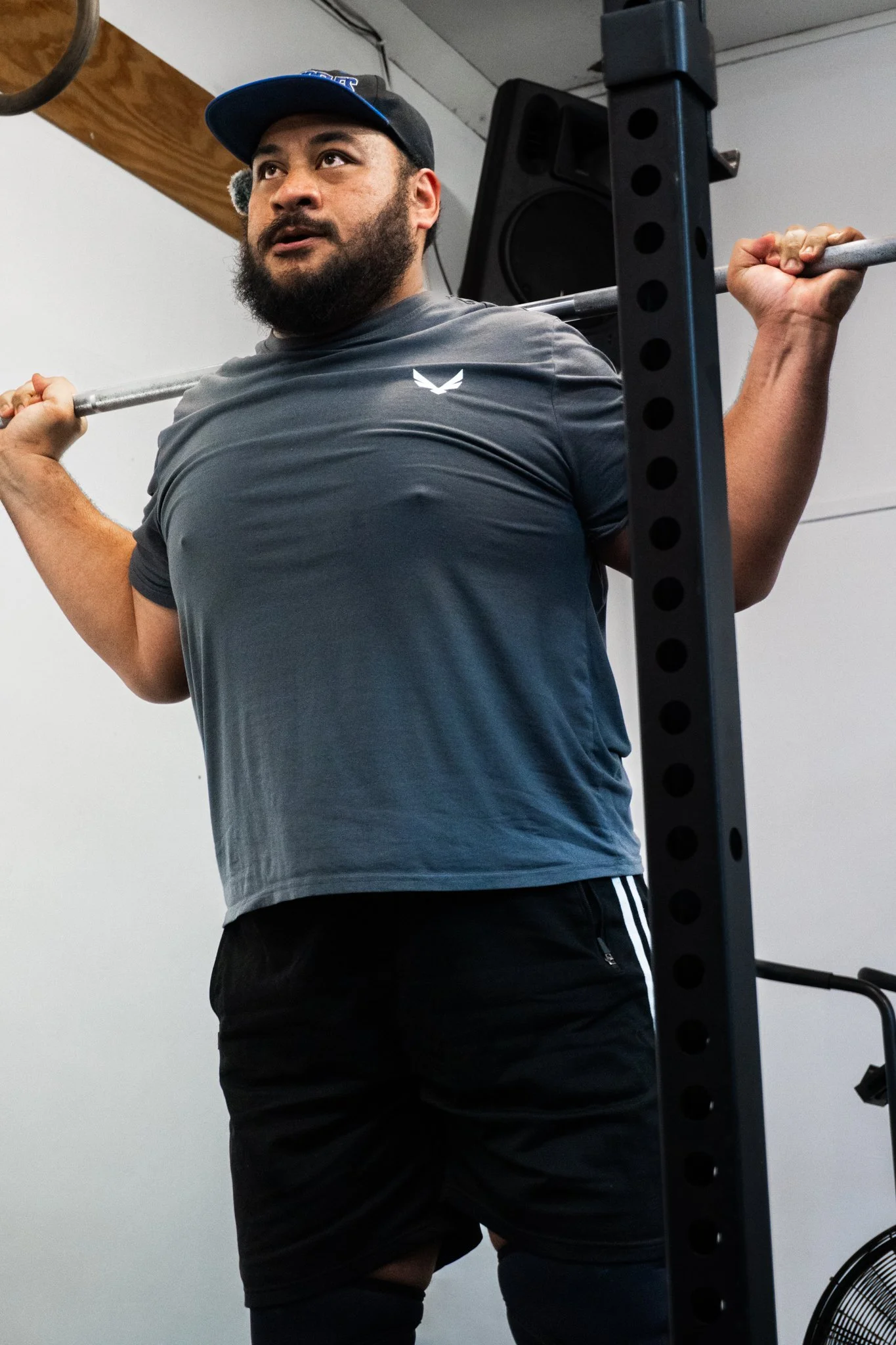A man doing a barbell squat in a gym, wearing a gray athletic shirt, black shorts, a cap, and knee sleeves.