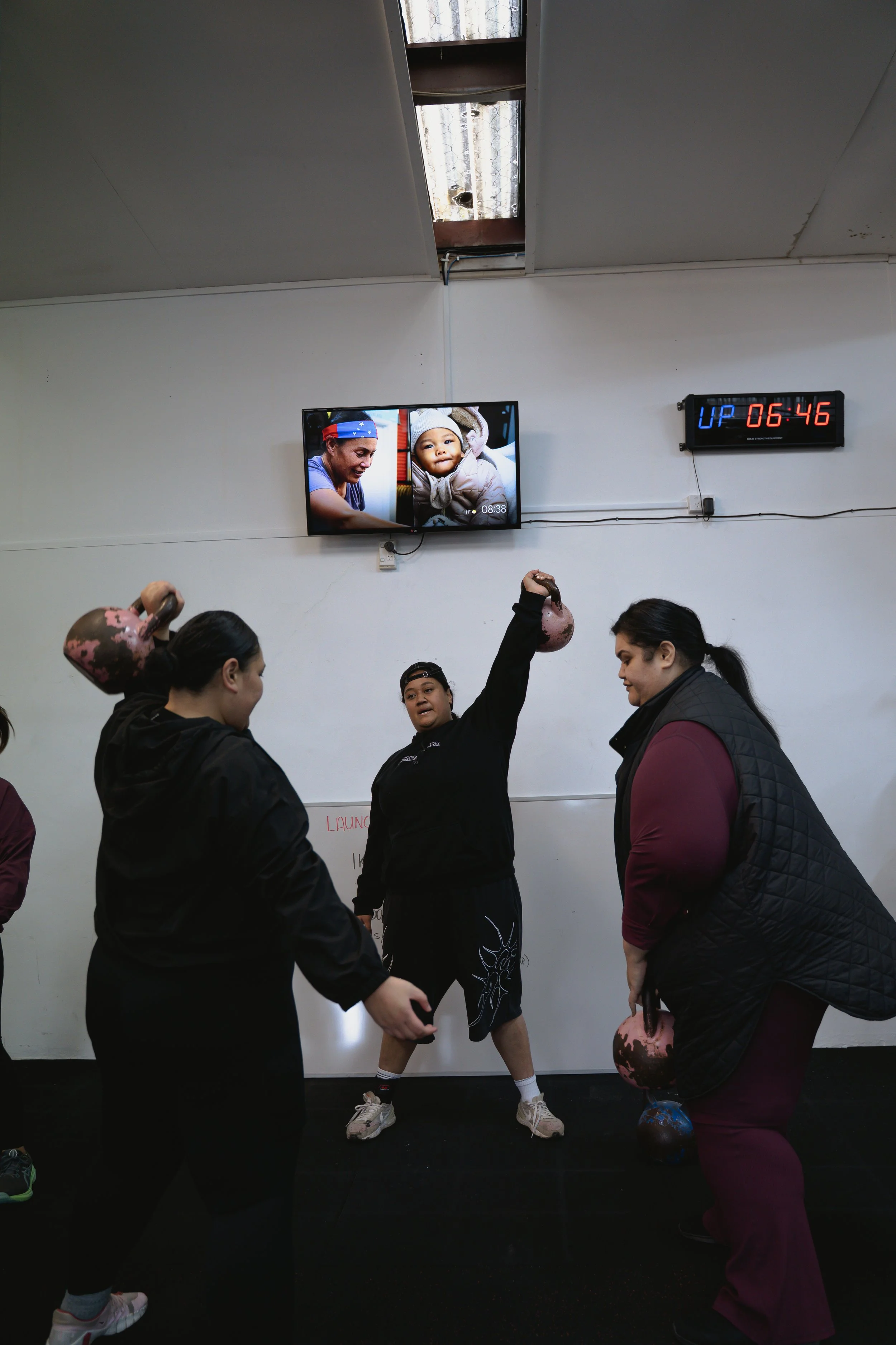 Women and a girl lifting kettlebells during a workout session in a gym. A digital clock on the wall shows 6:46, and a television monitor displays images of a woman and a child.