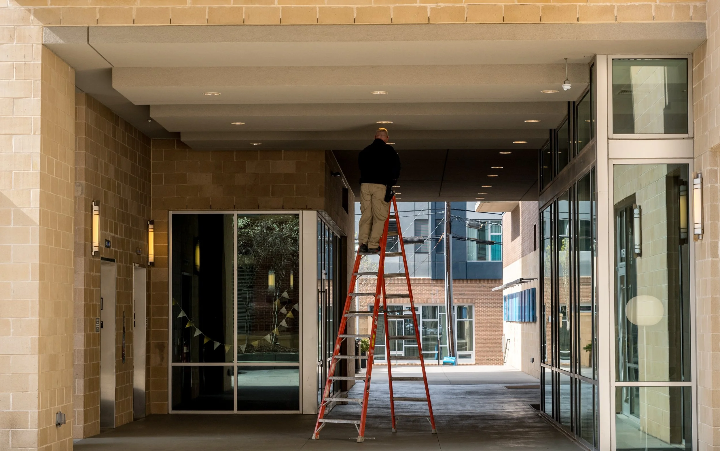 A man standing on a ladder, working on the ceiling of a building entranceway with glass doors and brick walls.