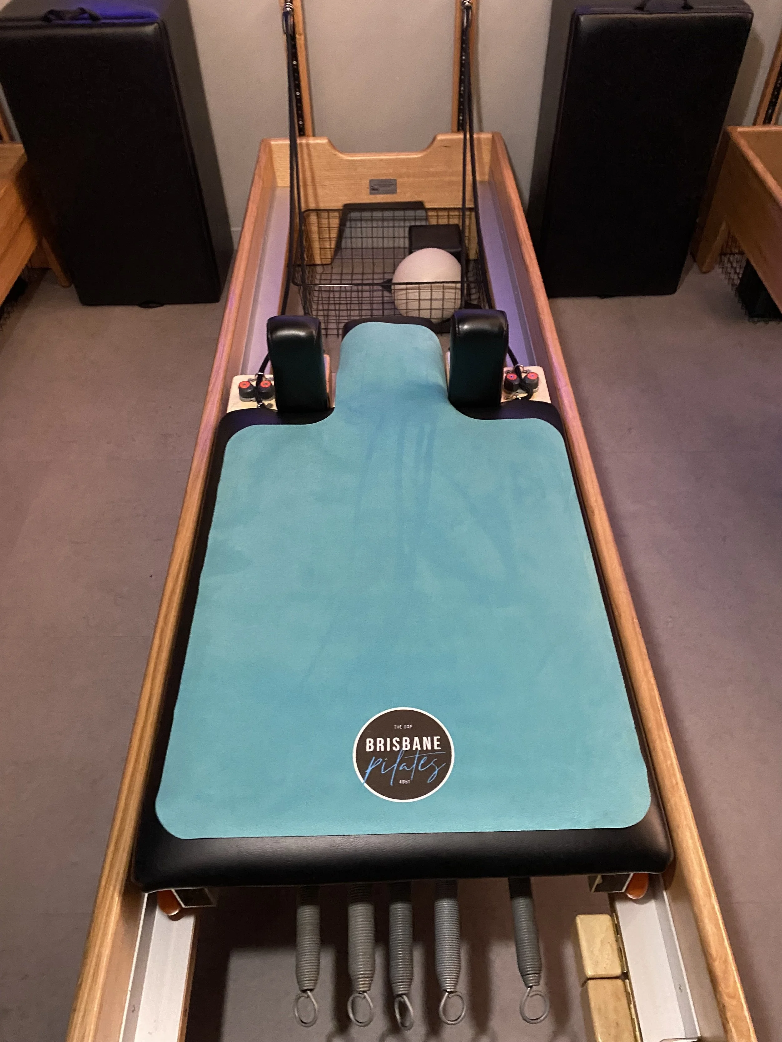 A shuffleboard table with a blue playing surface and a black border, featuring a circular sticker that reads 'Brisbane Pikes.' The table has two black paddles, a beige cue ball, and a small black object at the far end. The table is flanked by two large black speakers.