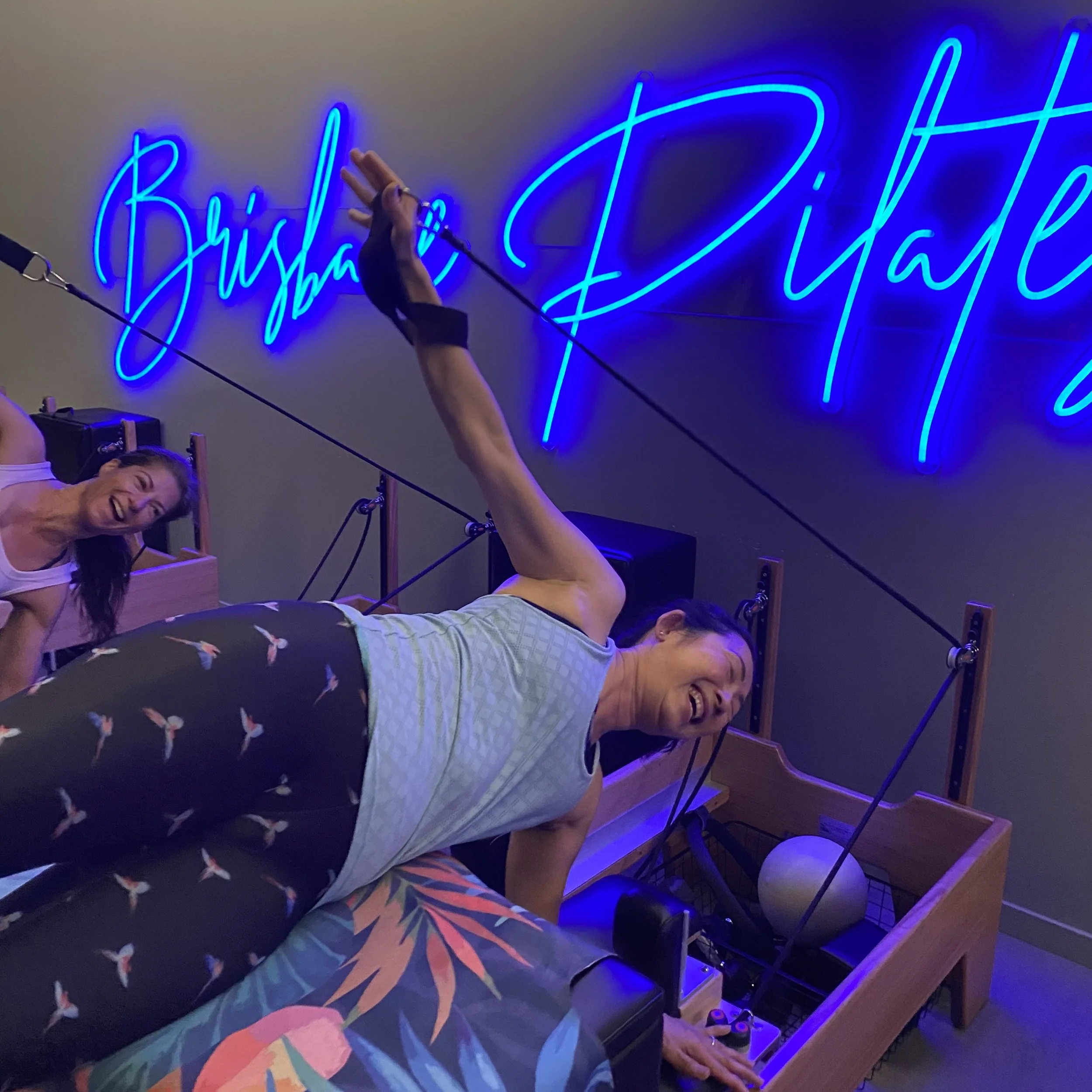 Two women exercising with Pilates reformer machines in a fitness studio with a neon blue sign saying 'Bring your life' on the wall.
