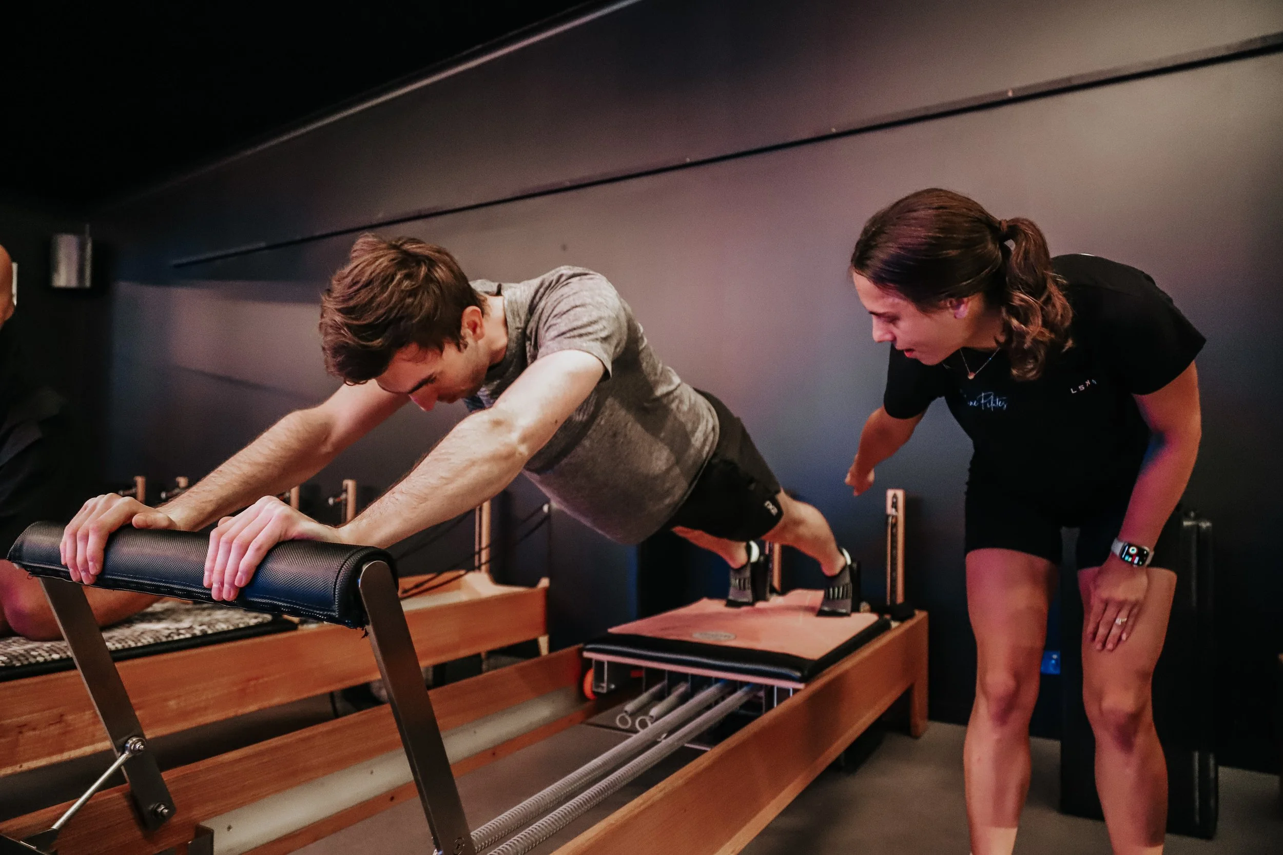 A man is doing a push-up on a fitness reformer machine while a woman, likely a trainer, is coaching him. The woman is standing to the side, leaning forward, pointing, and guiding him during the exercise.
