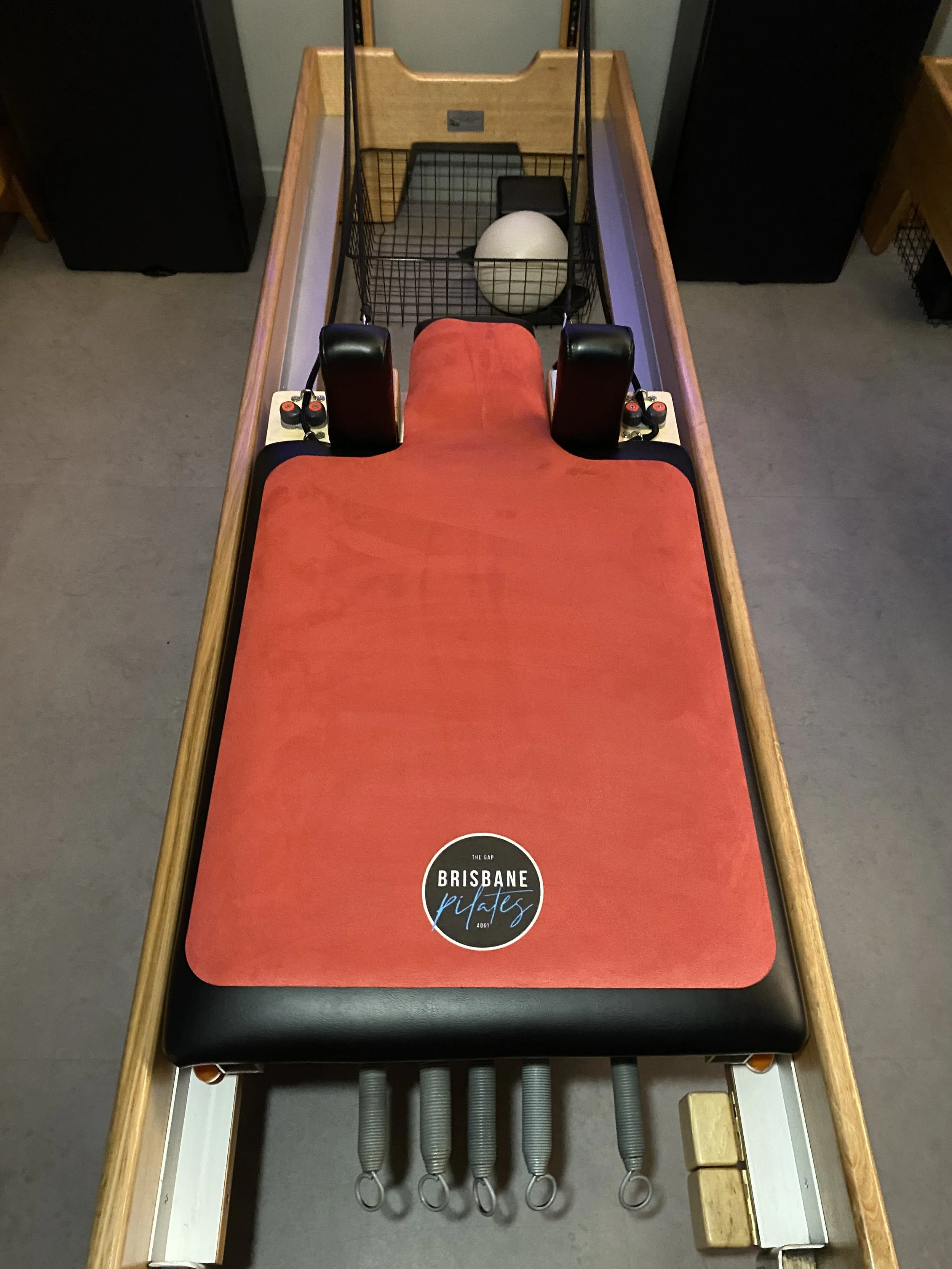 A pilates reformer machine with a red cushioned platform and black padding, located in a fitness studio. It has various springs attached at the foot and a sticker that says 'Brisbane Pilates.'