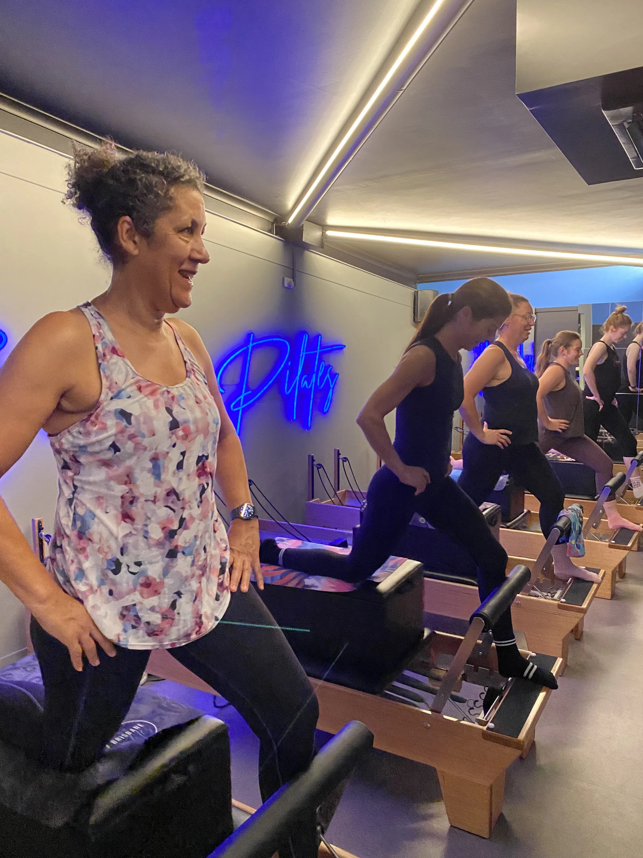 Group of women participating in a fitness class on reformer Pilates machines in a studio with neon sign reading 'Pilates' on the wall.