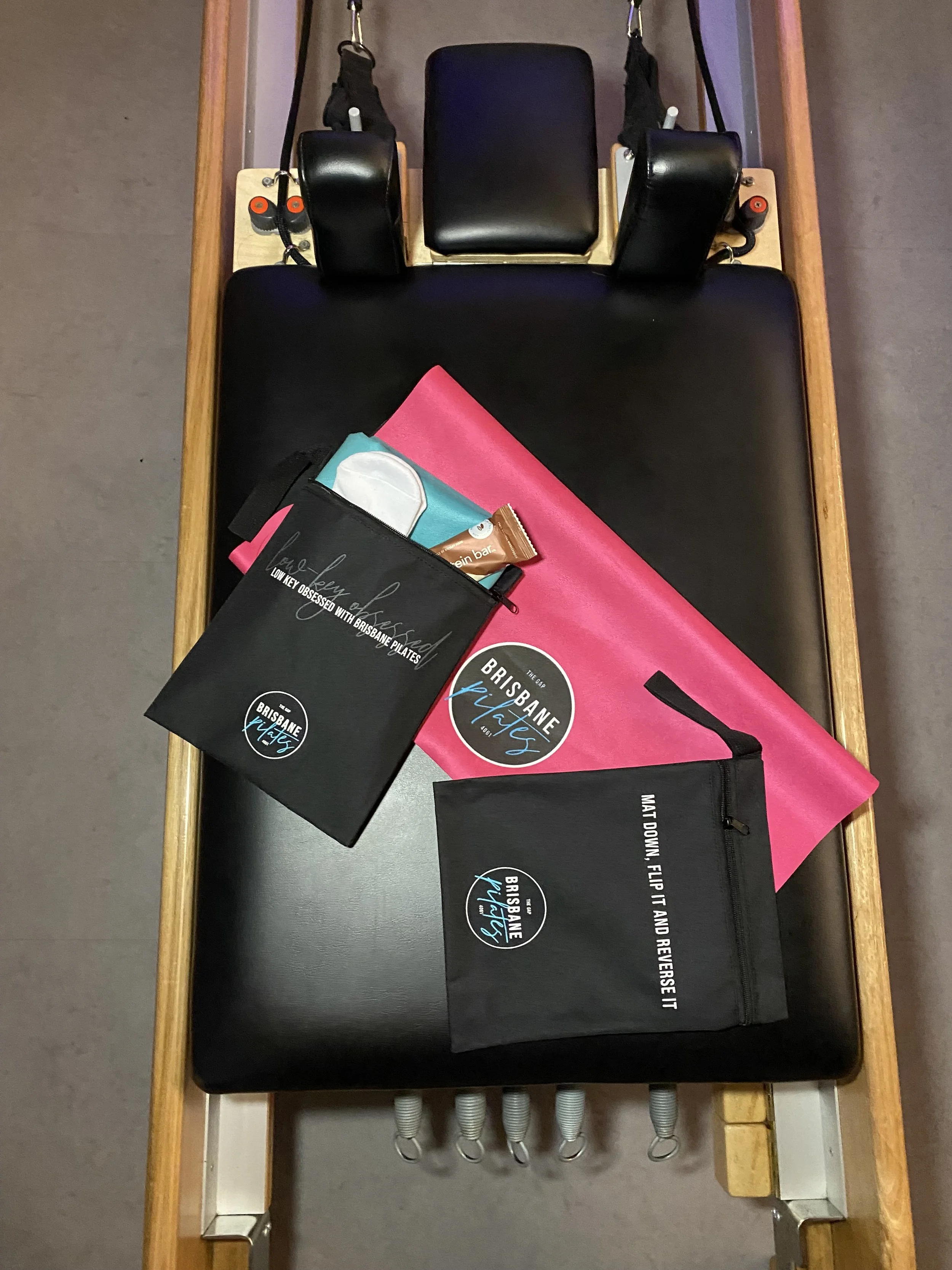 A Pilates reformer with black padding, wooden frame, and overhead straps. On the reformer, there is a pink exercise mat, a black bag from Brisbane Pilates with a logo, a small zippered pouch, a tube of skin care, and a cloth towel.