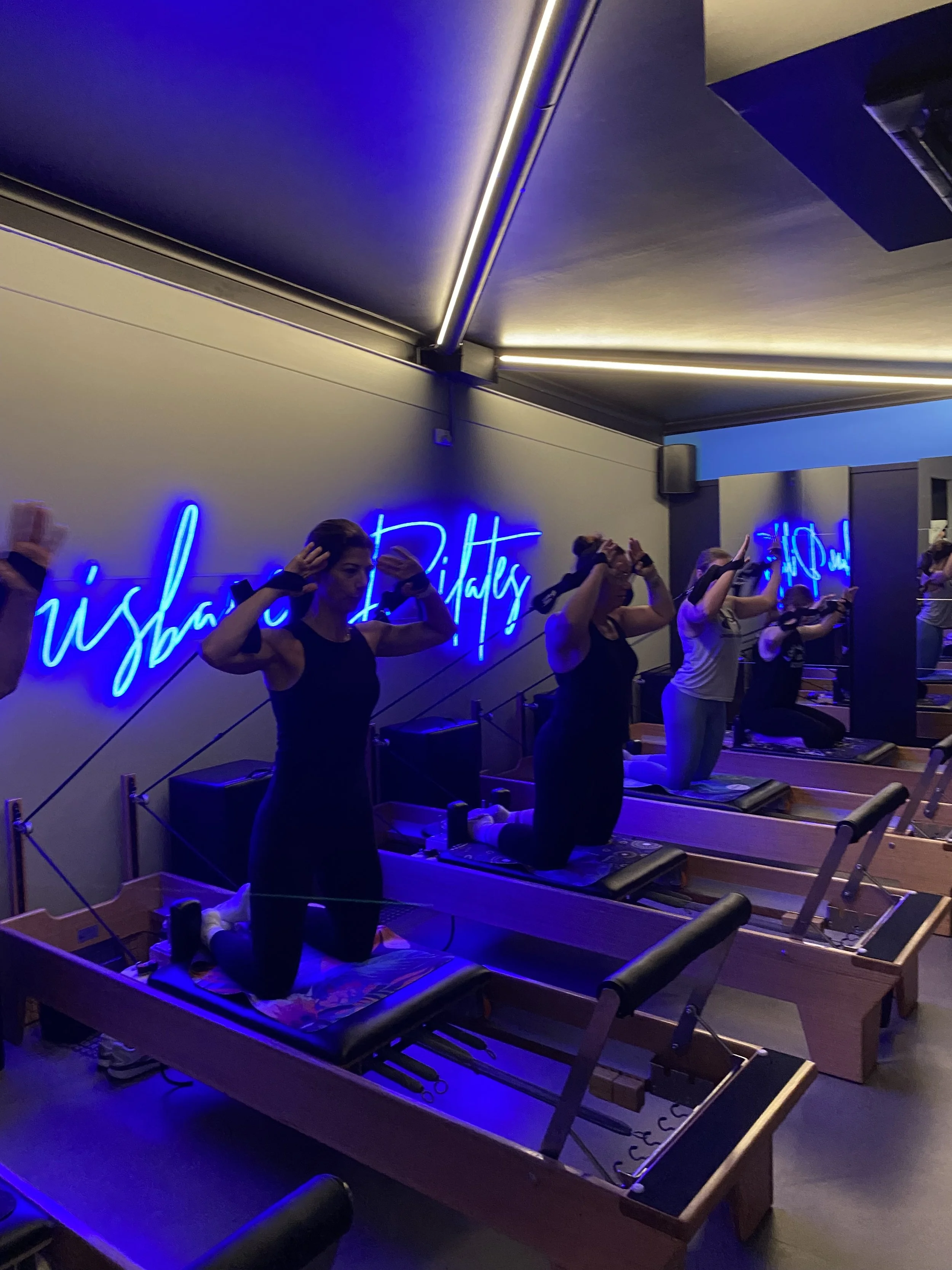 People participating in a Pilates class in a dimly lit room with blue neon lights and a sign that says 'Wisbach Pilates' on the wall, kneeling on Pilates reformer machines with their hands raised to their heads.