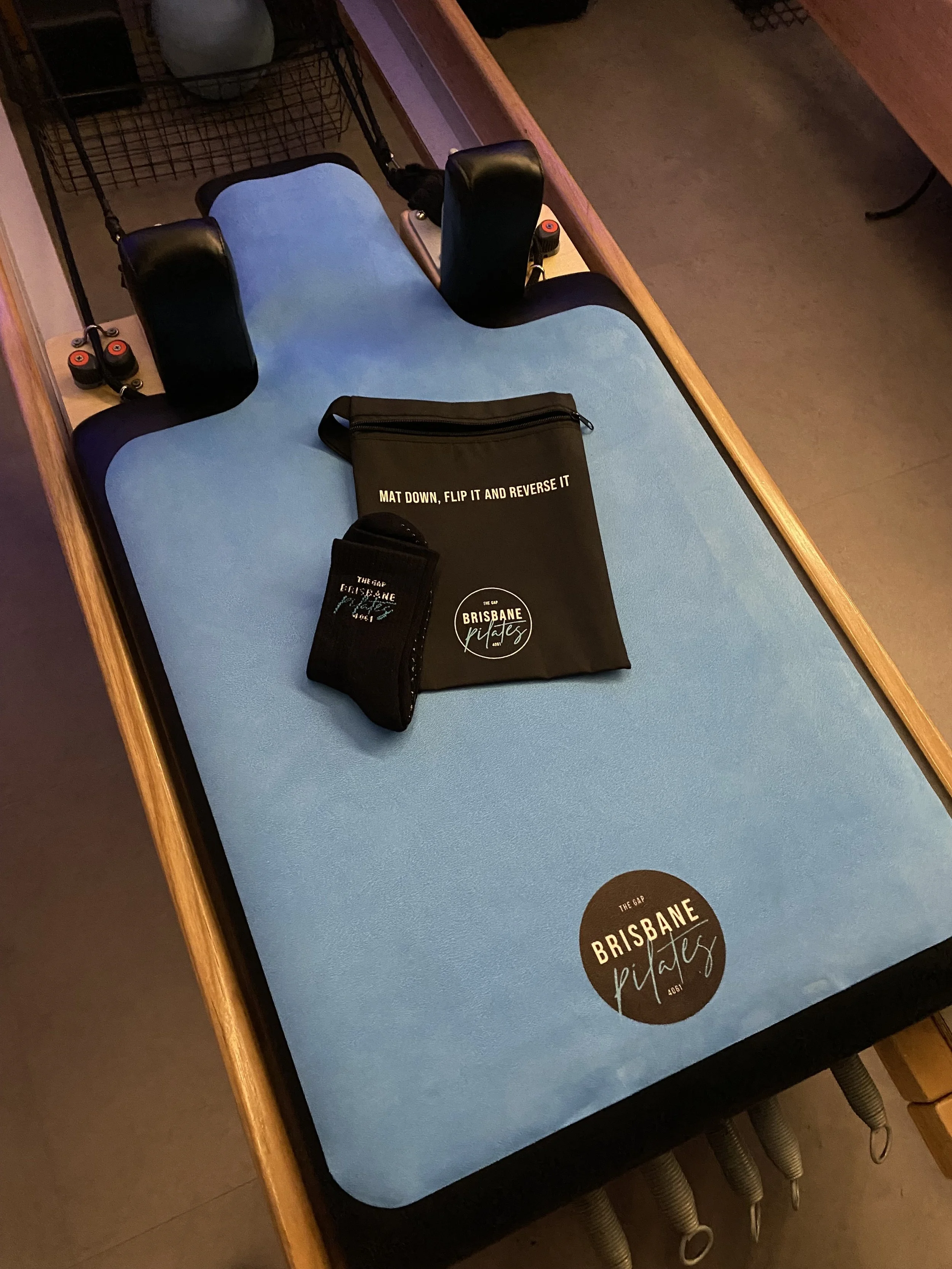 Foam-padded paddleboard on a wooden table with items on top, including a black zippered bag, a black towel, and a deck harness label reading 'Brisbane Kites'.