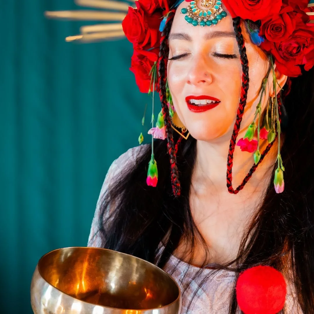 Woman holding a singing bowl, adorned with a floral headdress and closed eyes, in a meditative pose against a teal background.