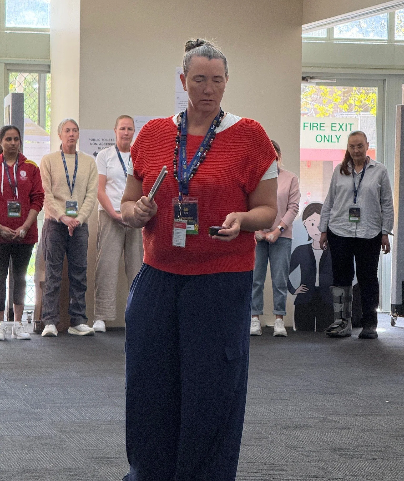 Group of people standing indoors, some wearing conference badges, with a woman in the foreground with a tuning fork in her hand and a hockey puck to activate for a tuning fork session.