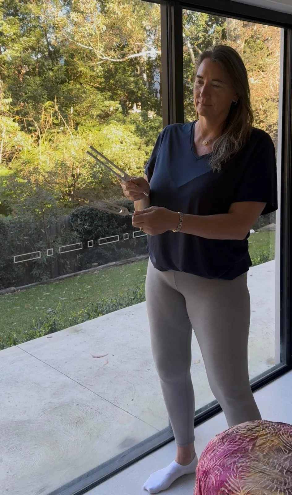 A woman standing inside near a glass sliding door, holding a tuning fork and a hockey puck, with a garden and trees visible outside.
