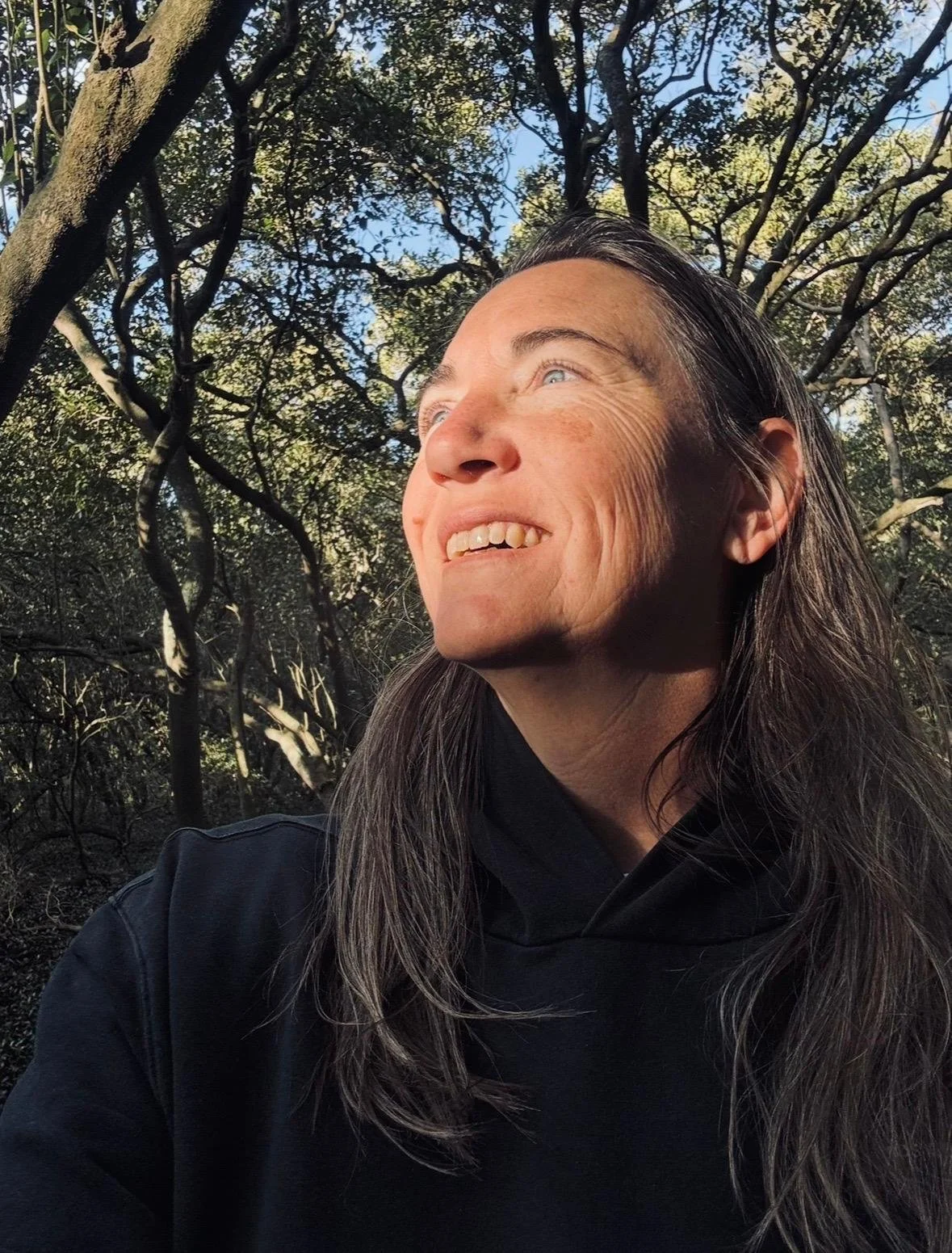 A woman with long gray hair and wearing a black jacket is outdoors, smiling and looking towards the sunlight with a background of trees and blue sky.