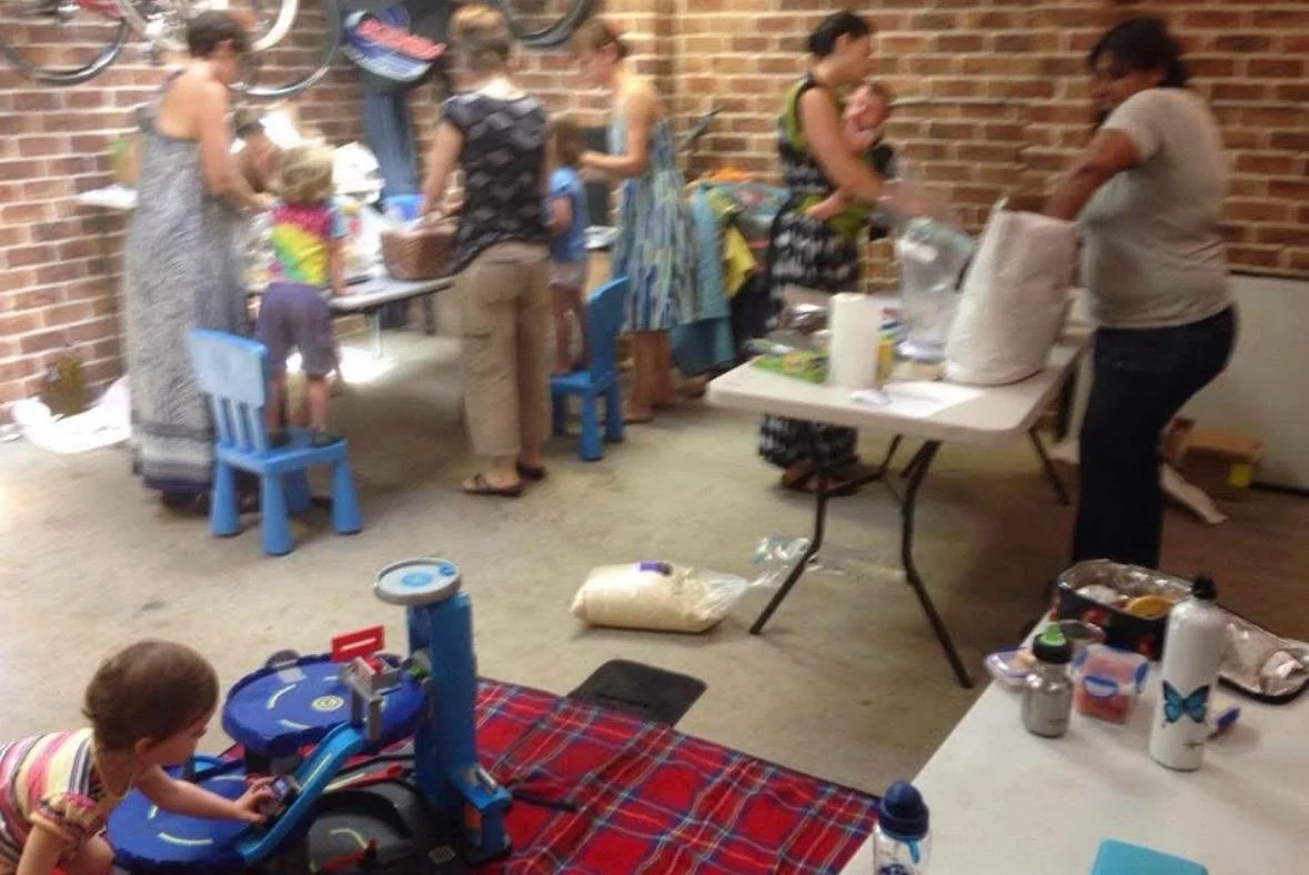 Multiple children and adults at a gathering with tables and supplies, inside a brick-walled room. Some people are serving food or drinks, while children sit and play.
