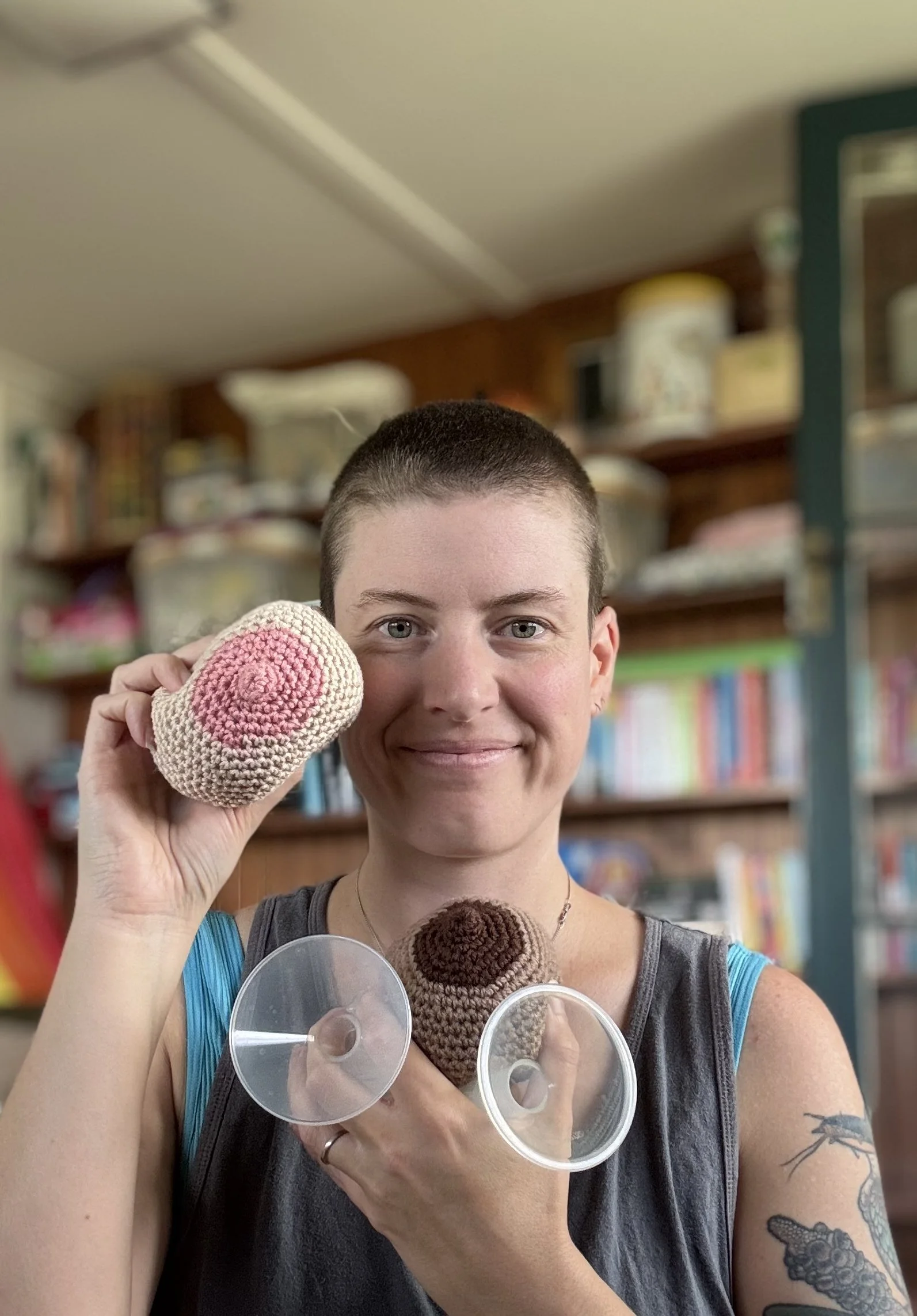 A white person with a shaved head smiling at the camera holding a crochet boob and some pump flanges.