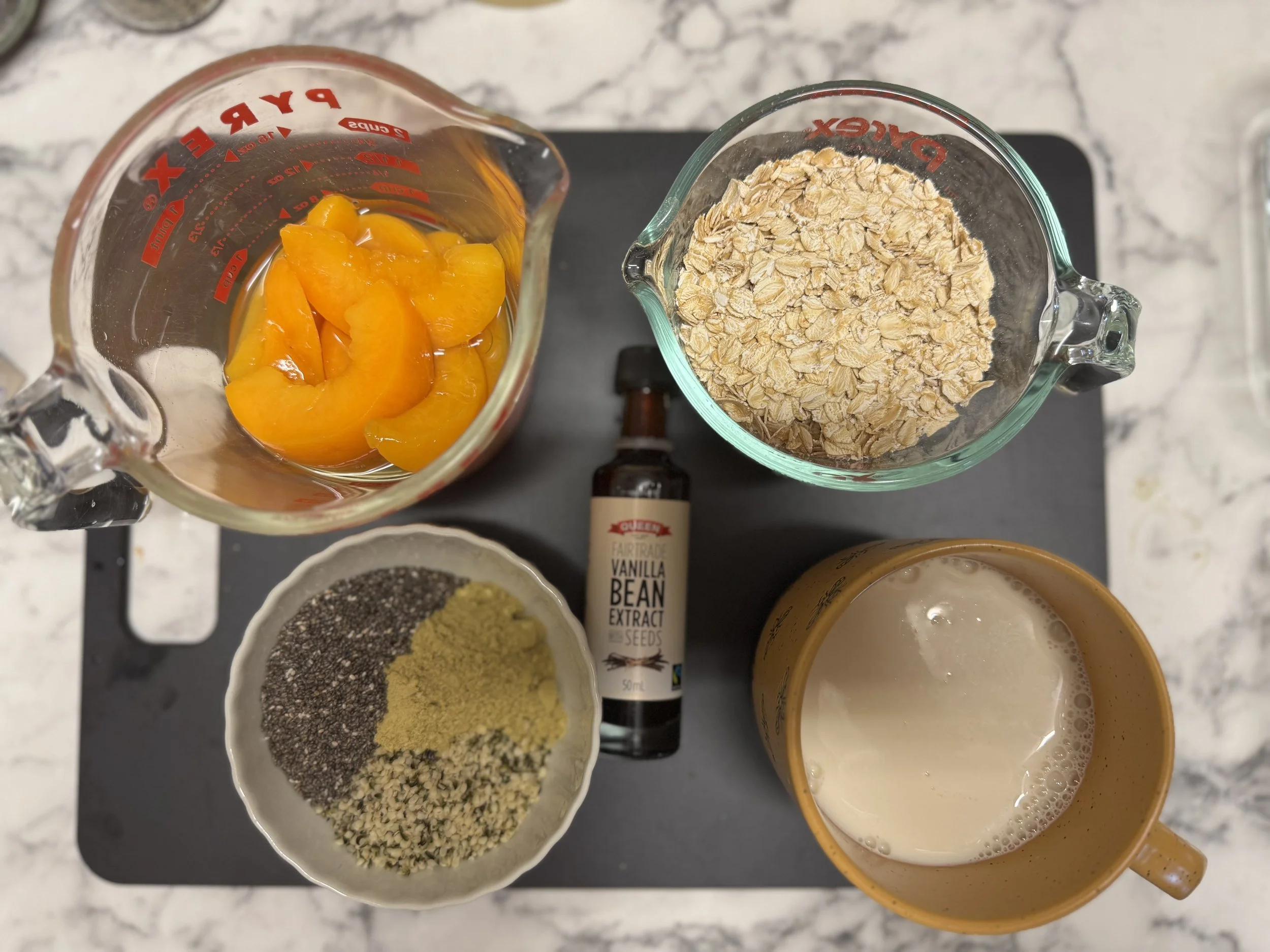 Ingredients for overnight oats: tinned peaches, rolles oats, chia, hemp, protein powder, vanilla, milk - all set out on a board.