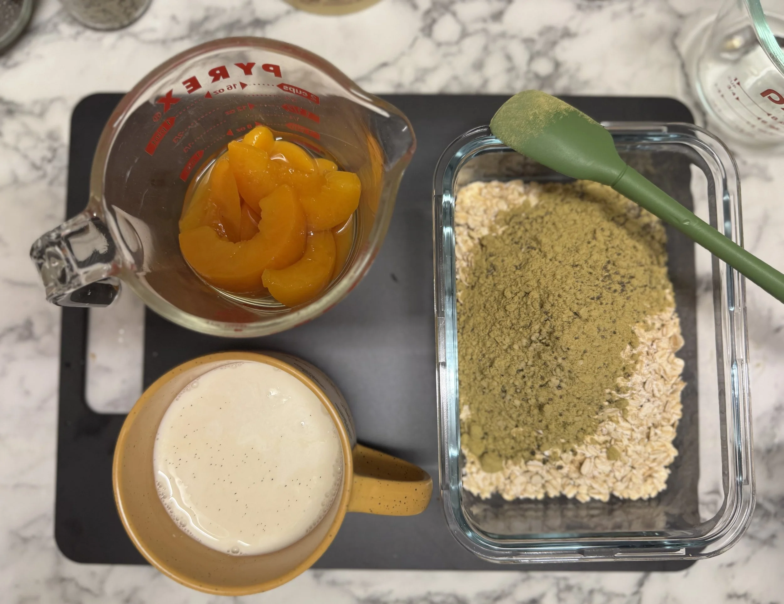 Oats, protein powder, chia and hemp seeds mixed in a glass container. Peaches, milk and vanilla still in jugs next to it.