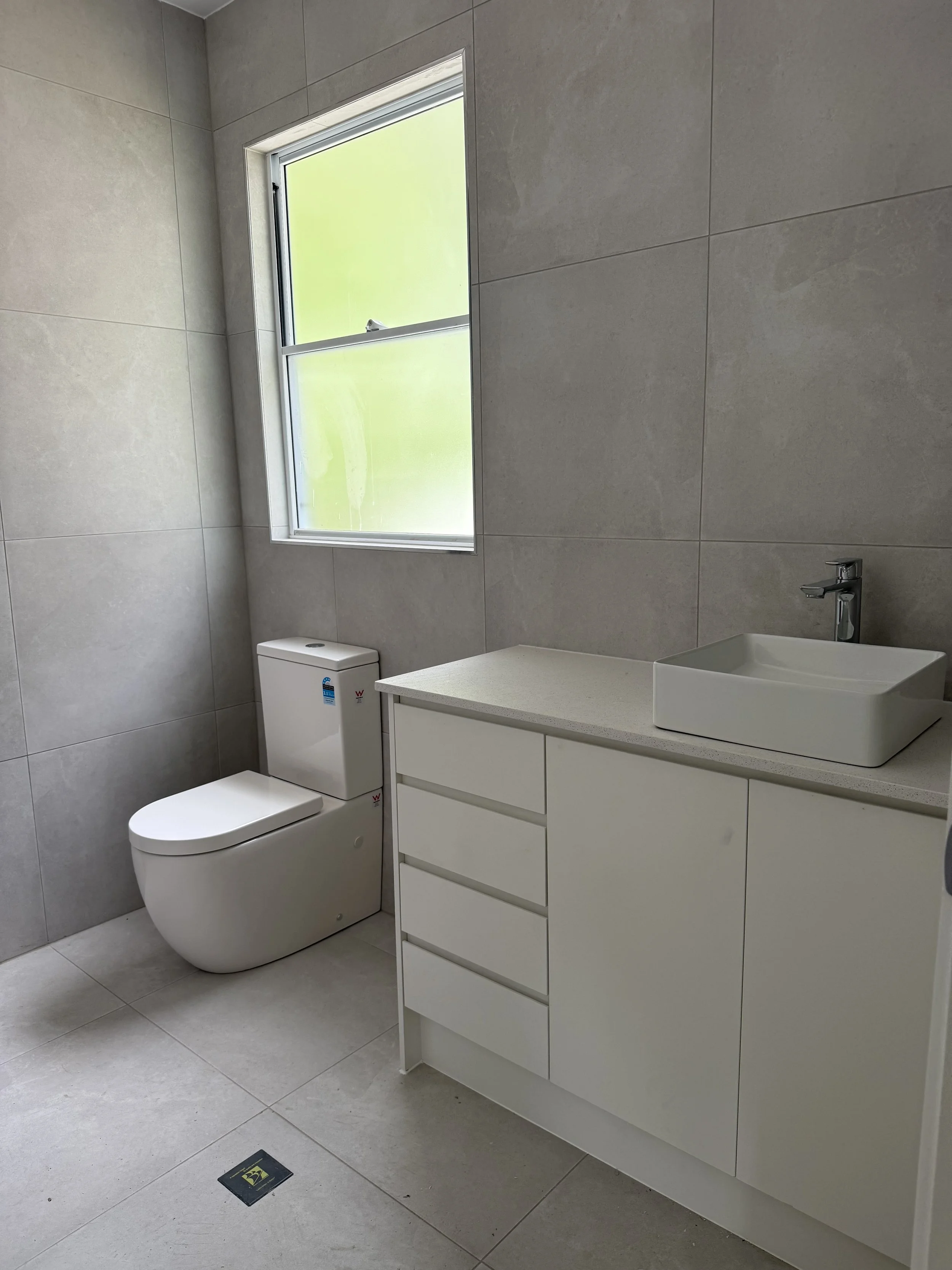 Modern bathroom with gray tiled walls and floor, a white toilet, a white vanity with multiple drawers, a square vessel sink, and a window with frosted glass.