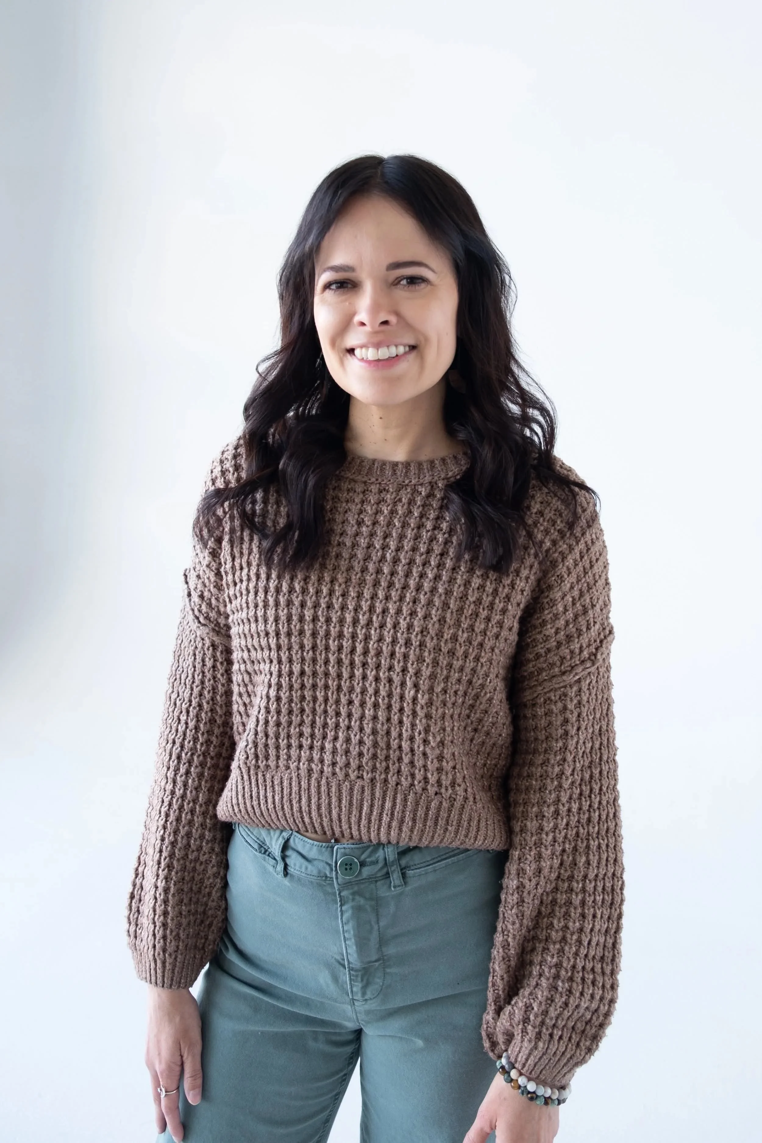 A woman with dark wavy hair wears a brown knitted sweater and gray pants, smiling at the camera against a plain white background.