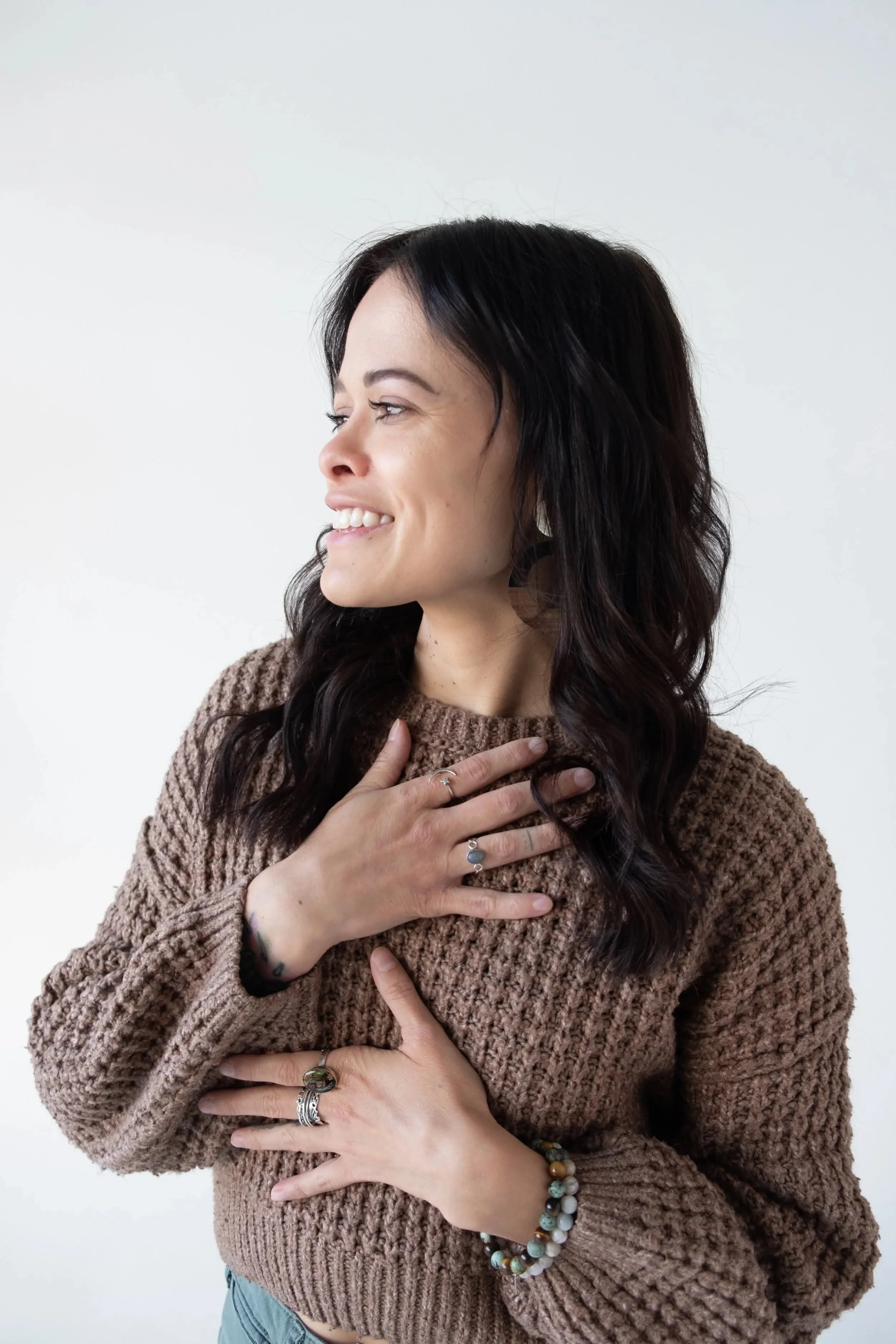 A woman with dark, wavy hair and light skin is smiling and looking to the side, wearing a brown knit sweater and multiple rings and bracelets, with her hand placed on her chest and stomach.