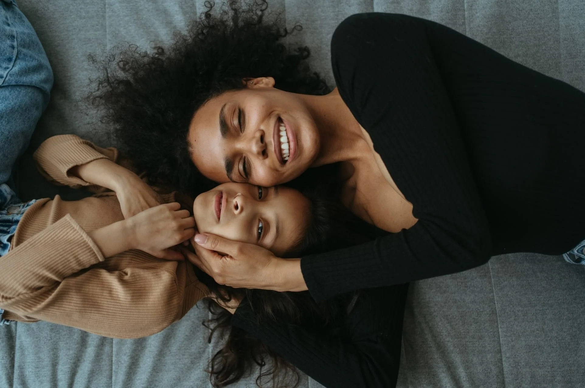 A woman with curly black hair and a black shirt lying on a gray couch, smiling with eyes closed, holding the face of a young girl with long dark hair, on a gray couch.