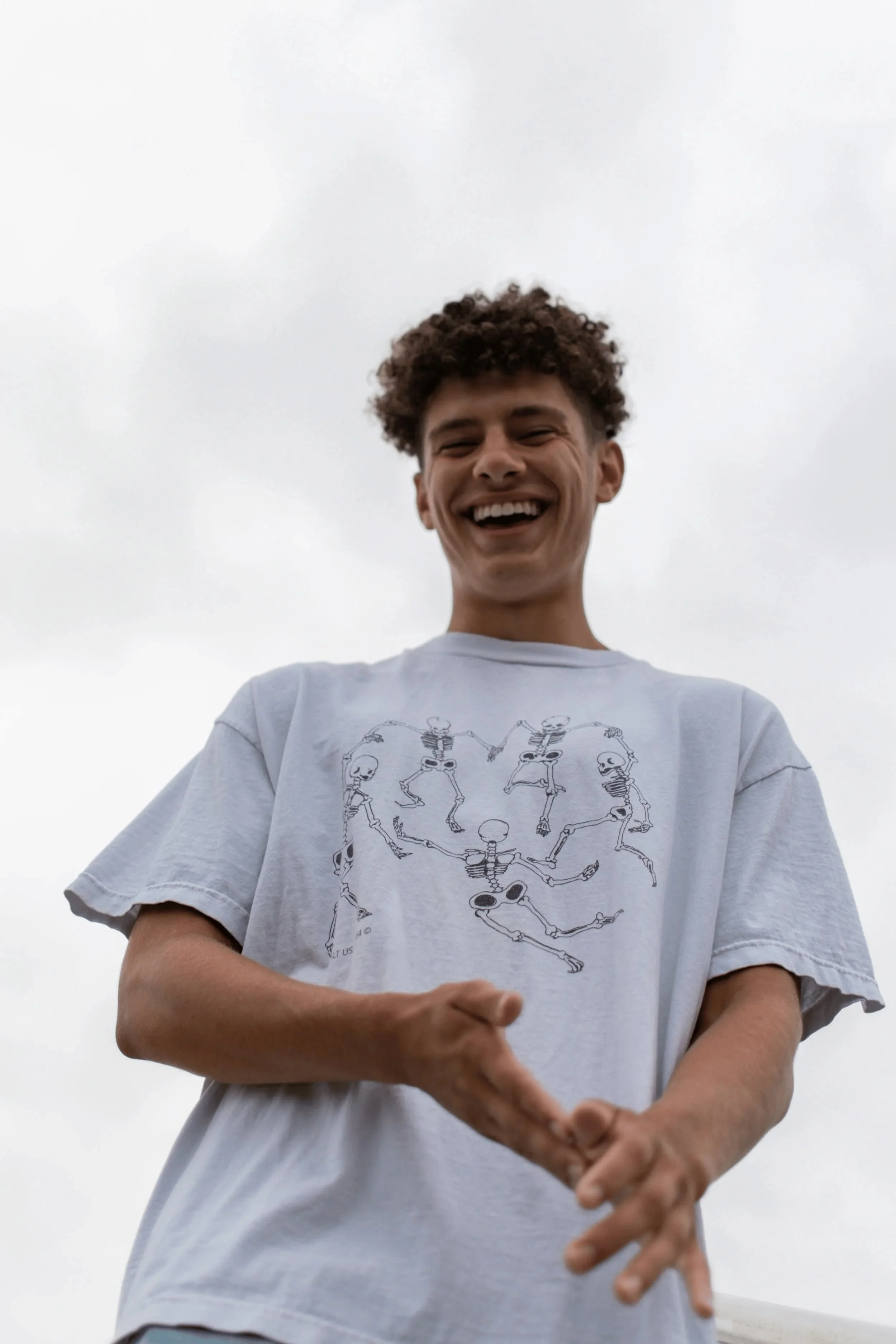 Young man smiling while looking down, wearing a white t-shirt with skeletal figures forming a circle, standing outdoors against a cloudy sky.