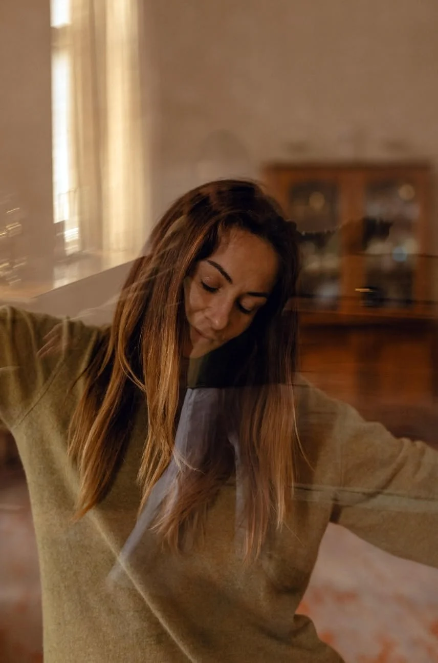 A woman with long brown hair and a beige sweater appears to be dancing or moving to music in a living room, with a blurred background and warm ambient lighting.