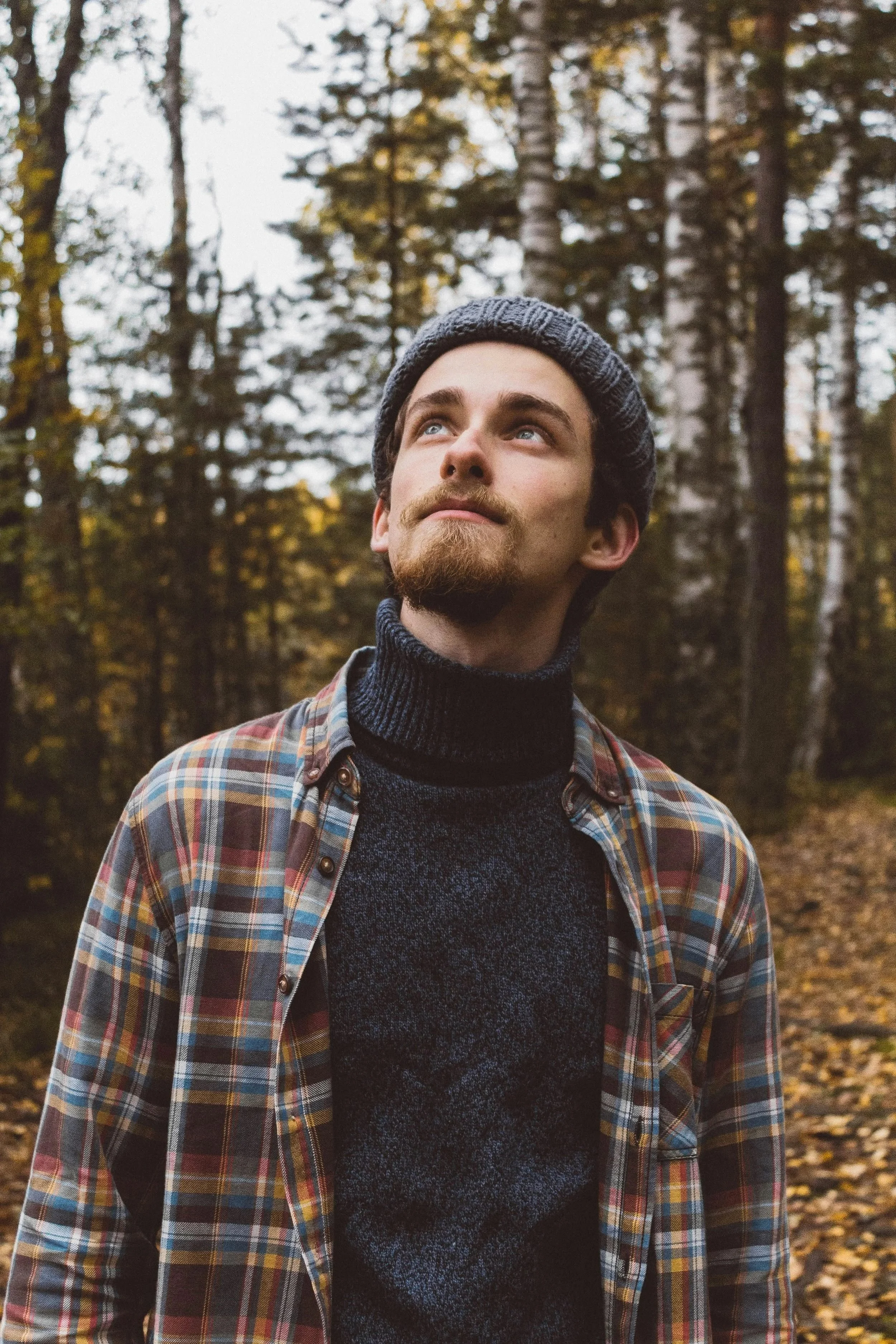 A young man with a beard and mustache wearing a dark knit beanie, a navy turtleneck sweater, and a plaid shirt outdoors in a wooded area during autumn.