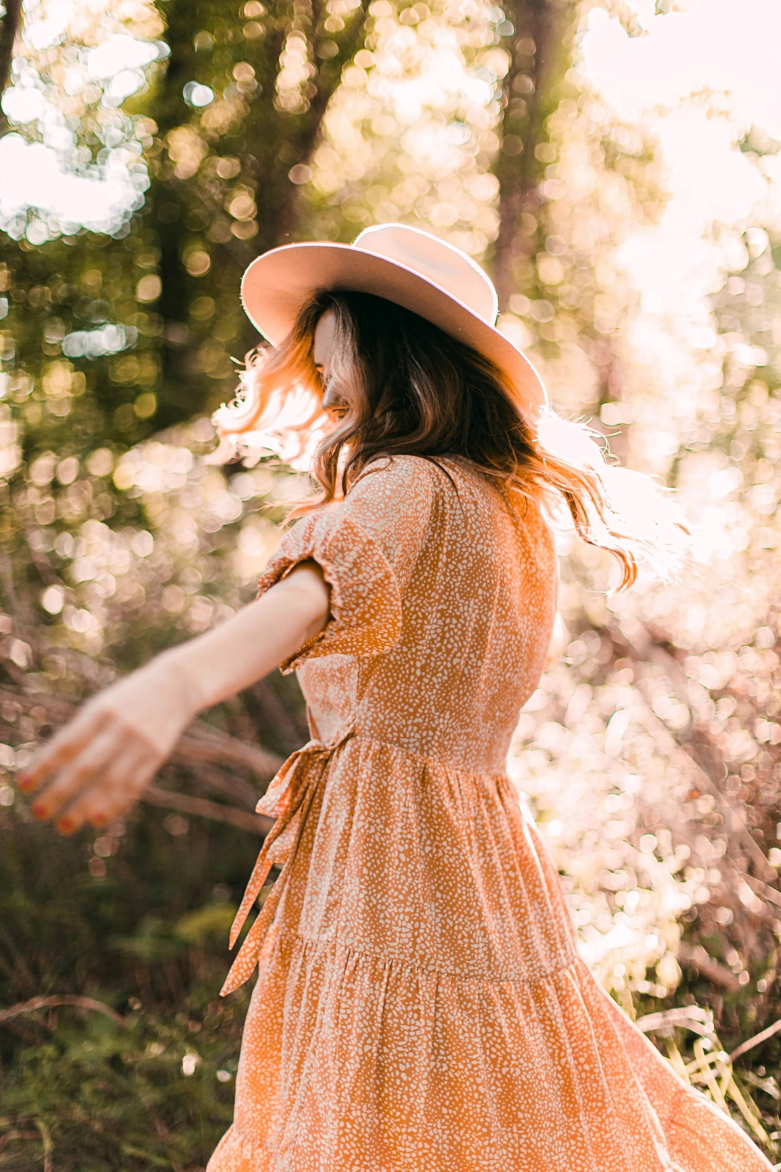 A woman with brown hair wearing an orange patterned dress and a large beige sunhat, standing outdoors in a forest with sunlight filtering through the trees.