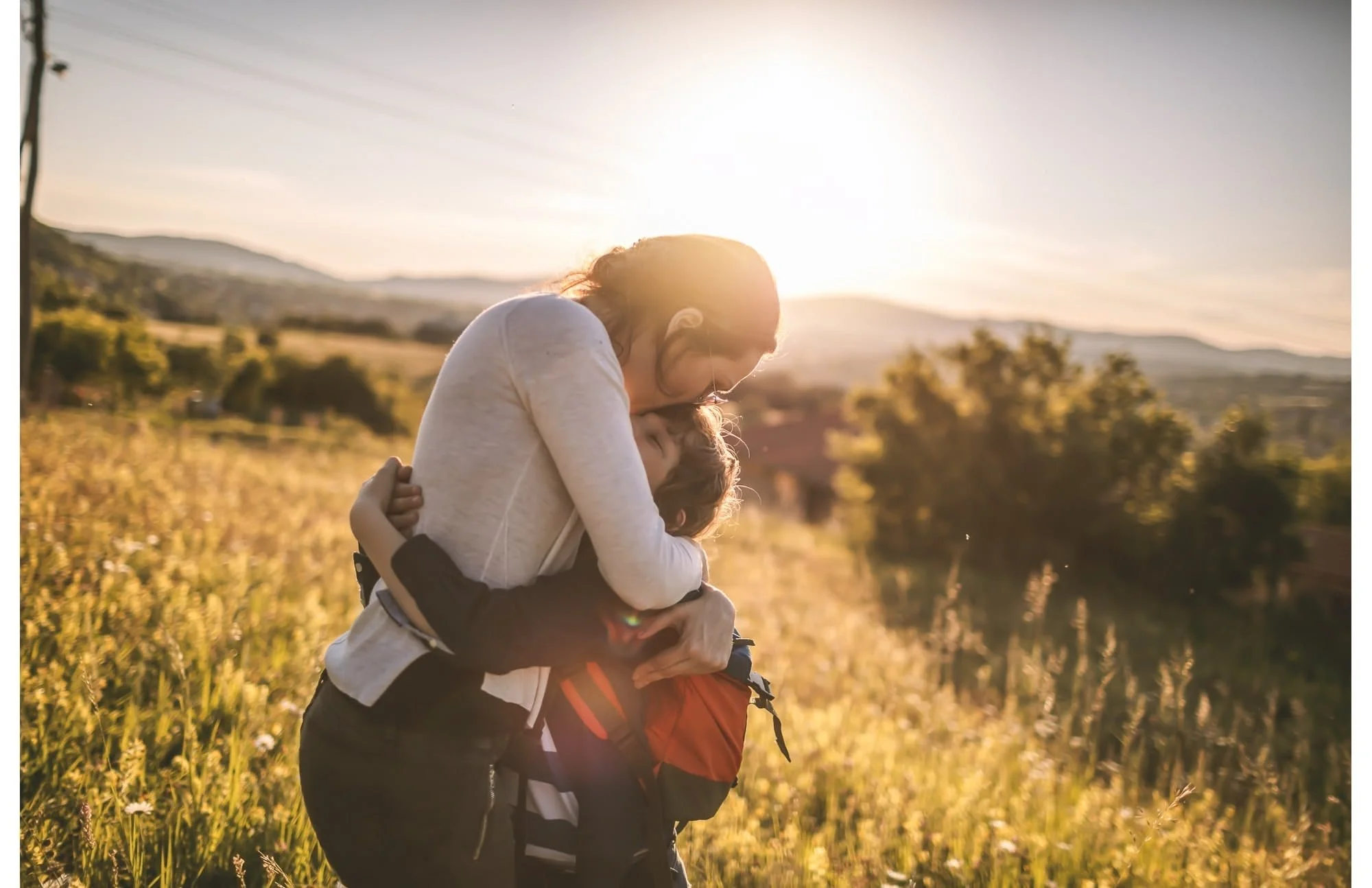 A woman hugging a child in a field during sunset with mountains and trees in the background.