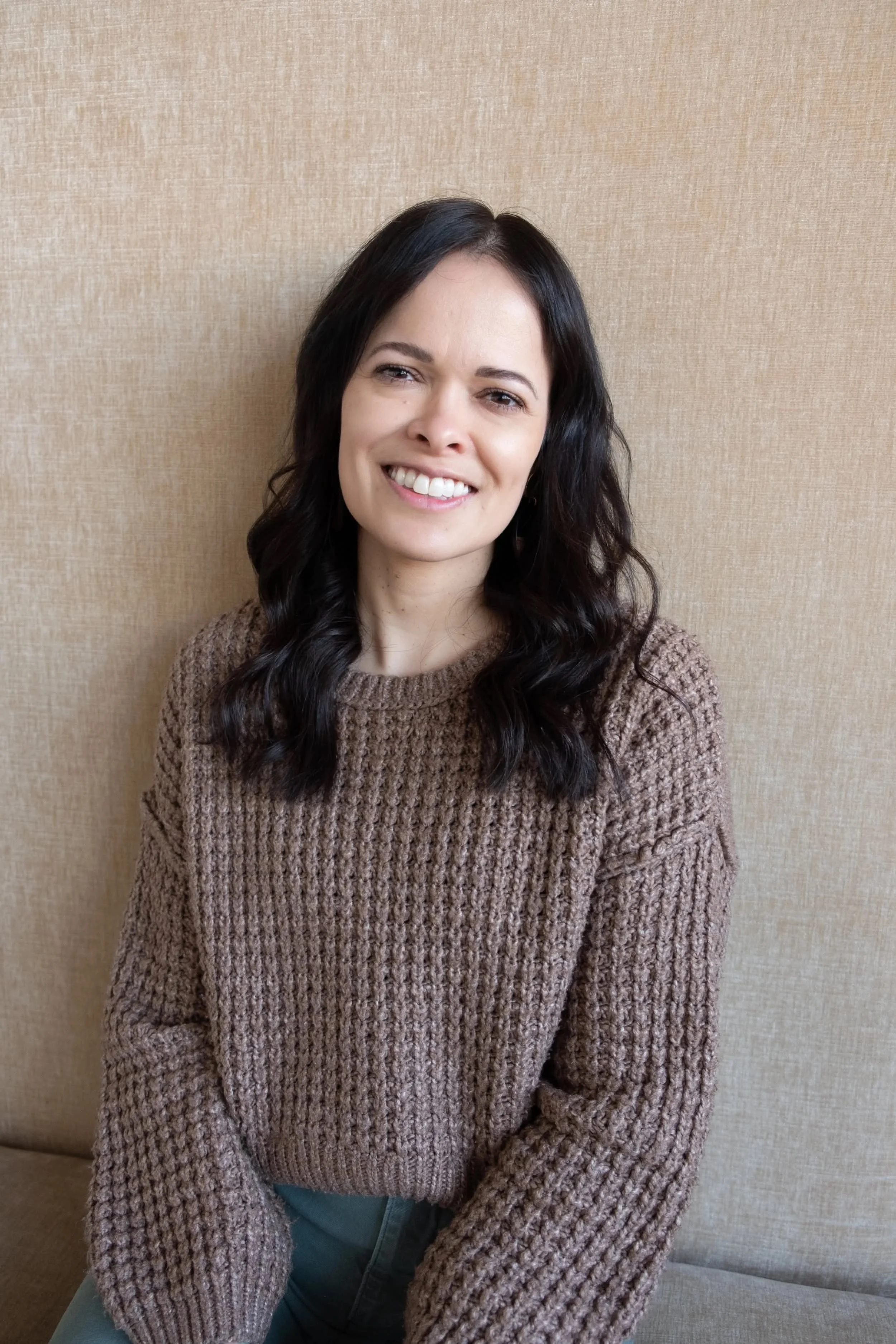A woman with dark wavy hair and a brown knitted sweater, sitting against a beige textured wall, smiling at the camera.