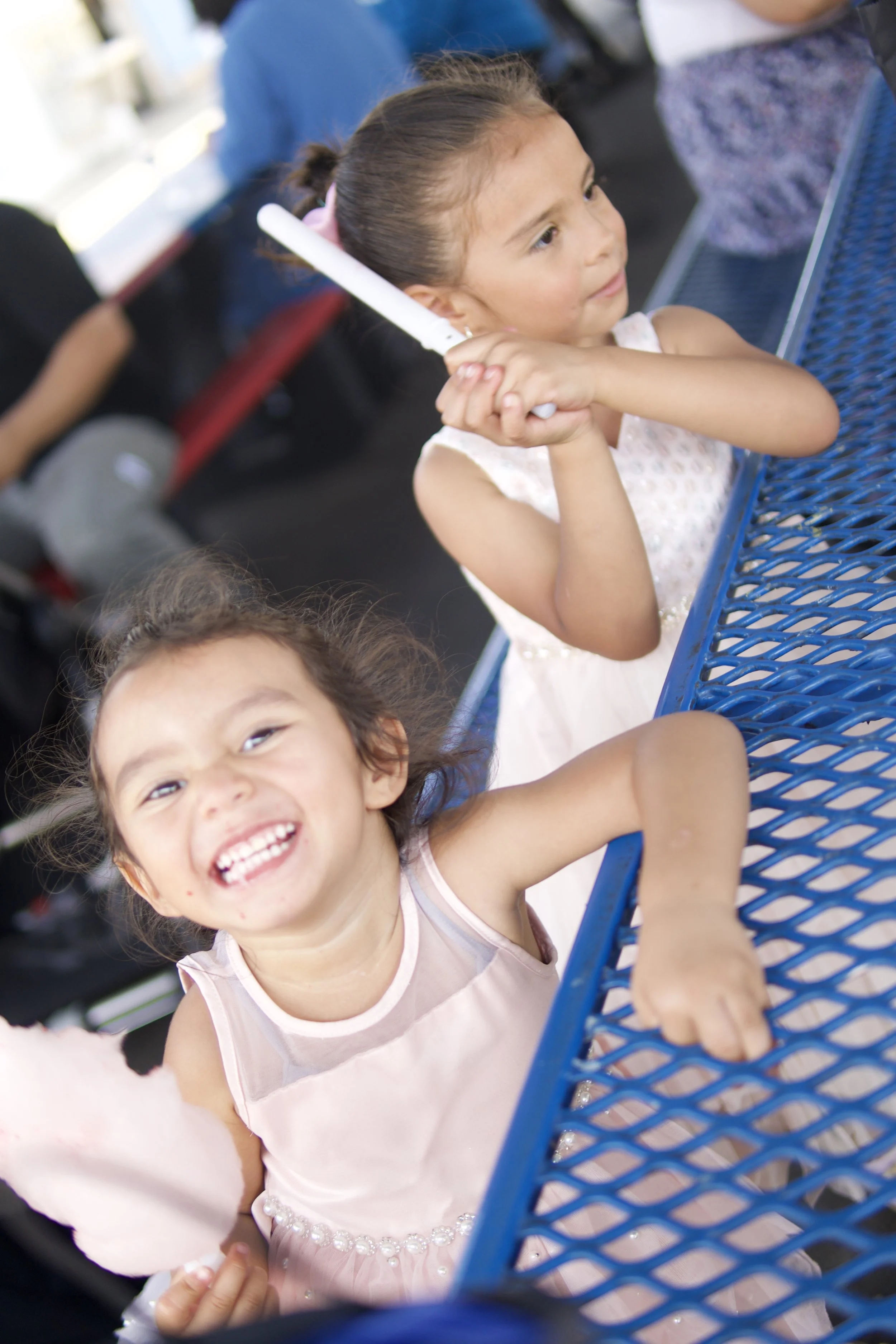 Two young girls sitting at a blue outdoor table, smiling and looking joyful. One girl has her hands clasped and is holding a white glow stick near her cheek, and the other girl is laughing and reaching to hold the first girl's hand.
