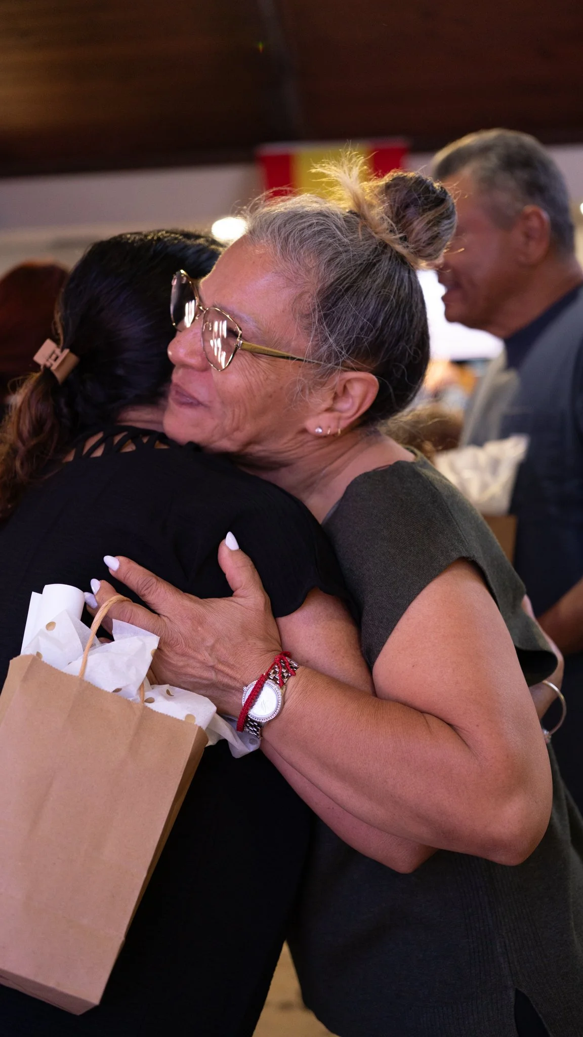 Two women hugging warmly, with one holding gift bags, in a social setting.