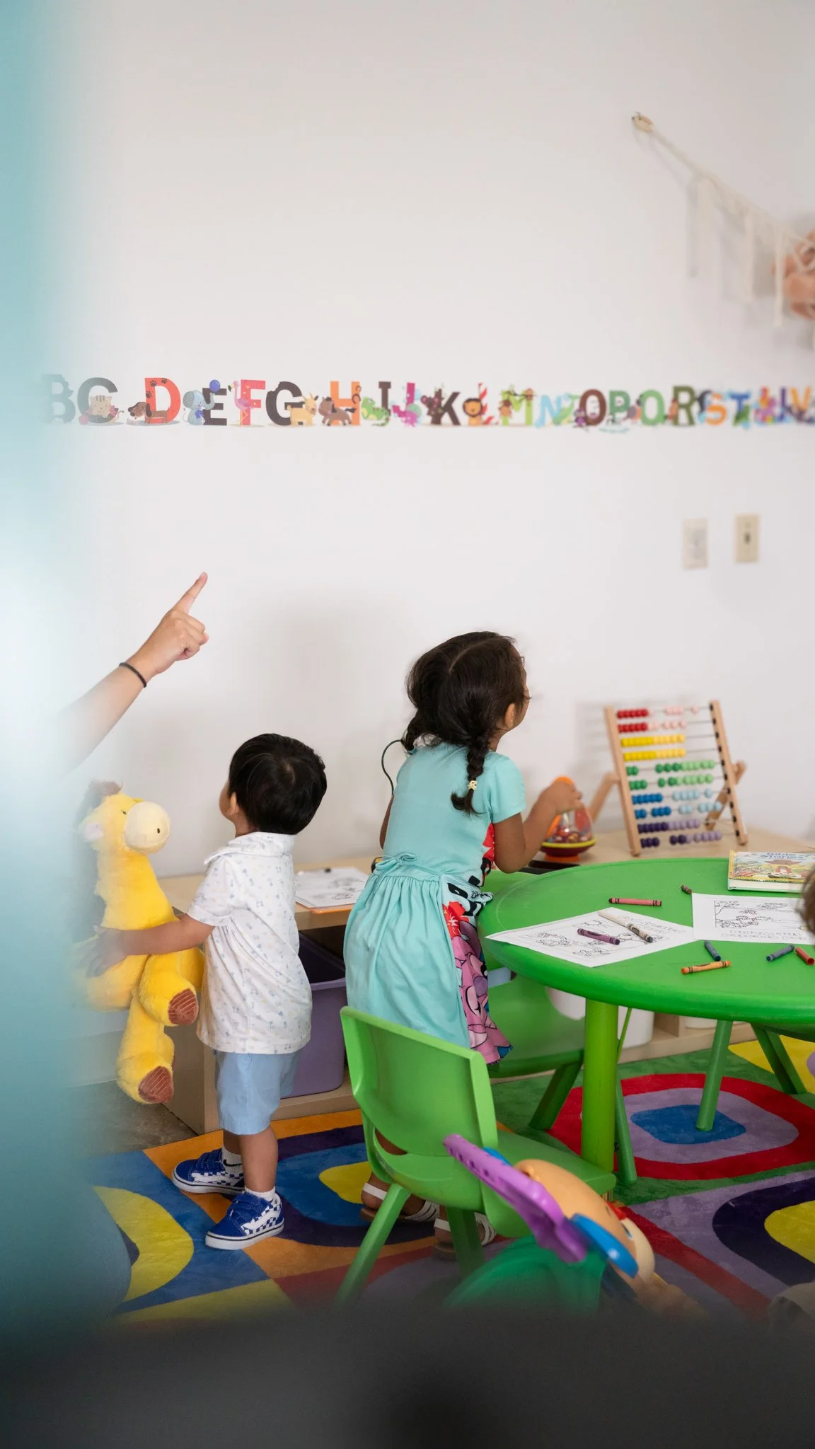 Children in a colorful classroom with alphabet decorations on the wall, including a girl and a boy playing with toys at a green table.