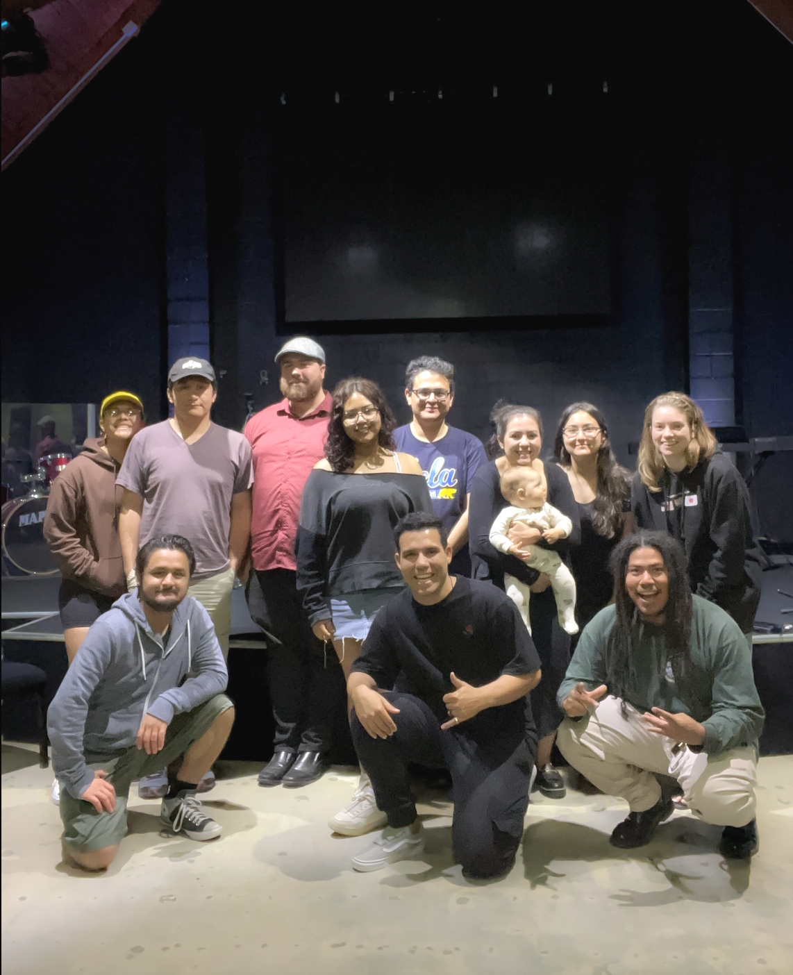 Group of people posing for a photo in a dark room with a large screen and musical instruments in the background.