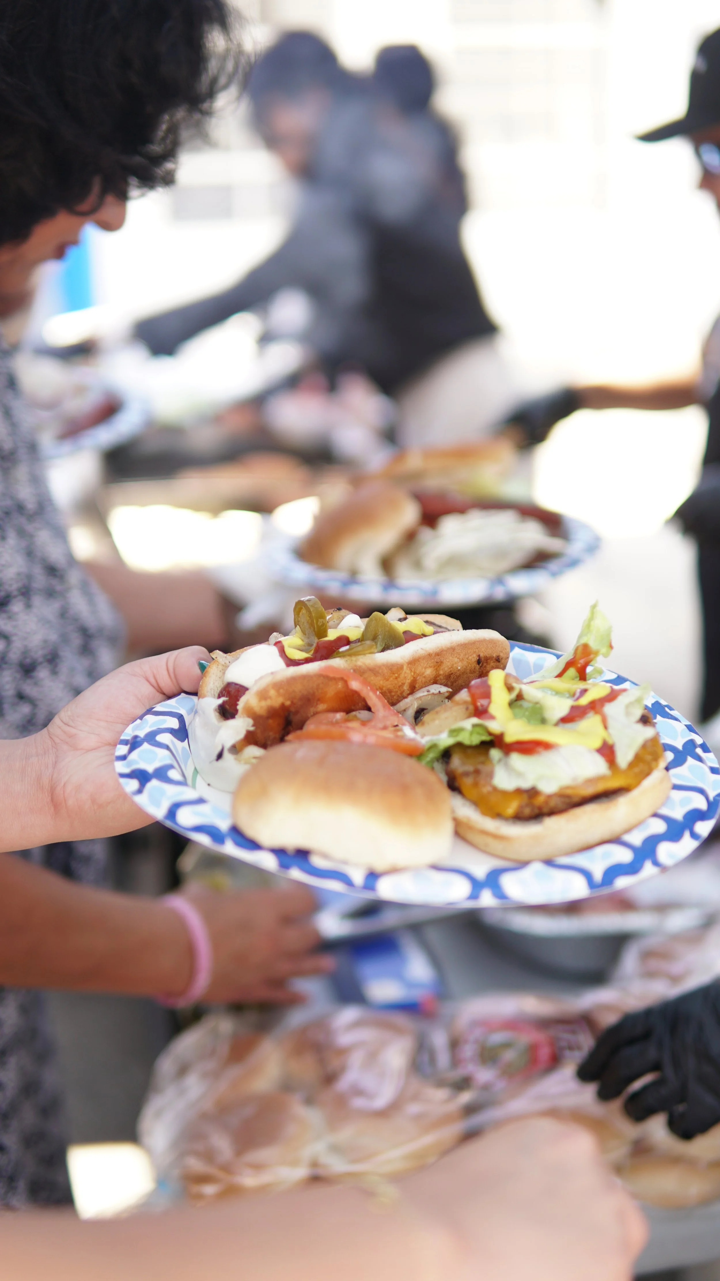 Assorted hot dogs with various toppings on a disposable plate being handed to a person at a buffet or food service line.