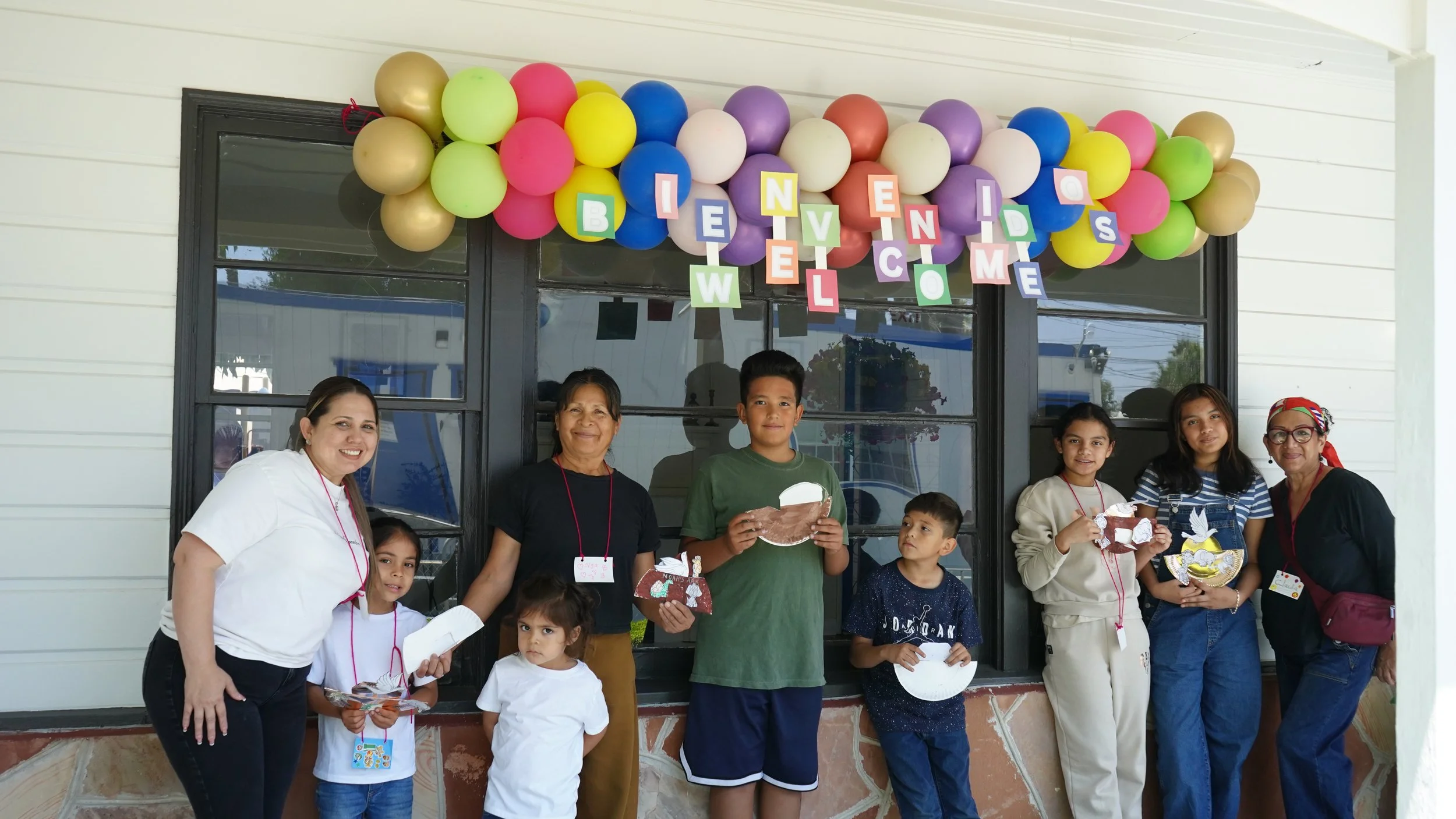 Group of children and adults celebrating at a birthday party, standing in front of a decorated wall with balloons and a welcome sign. Some kids hold party favors and cakes.